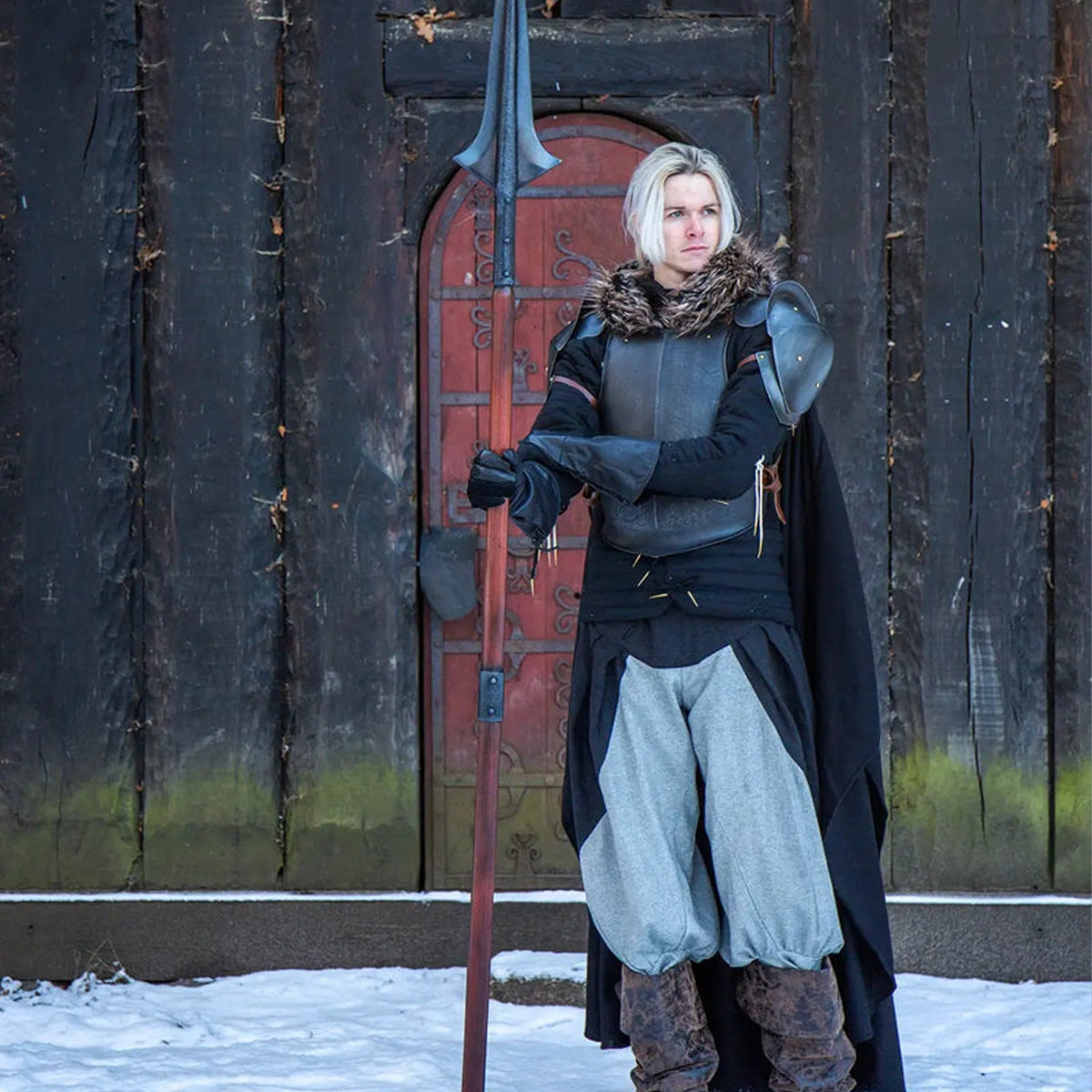 Person in armor with a spear standing in front of a wooden door in a snowy setting, highlighting compact gear.