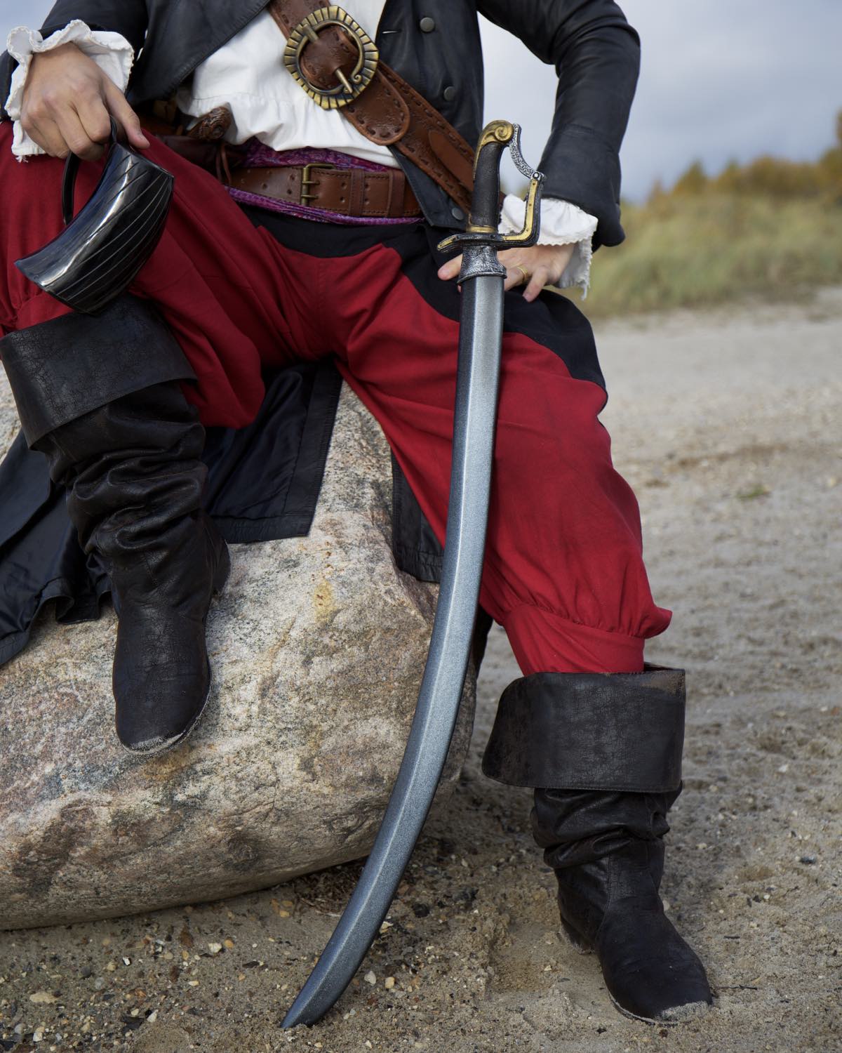 A pirate resting on a rock with a curved sword, showcasing vibrant red pants and black boots.