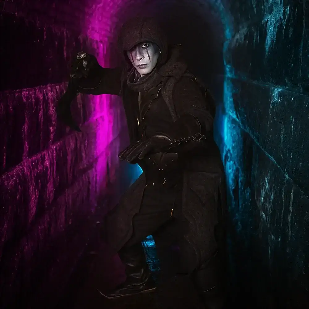A mysterious figure in a hooded cloak poses dramatically in a dimly lit tunnel with colorful lights.