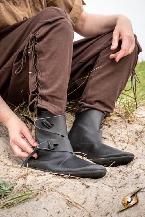 Shoes Jorik: Black leather Viking footwear worn by a person sitting on sand with brown pants.