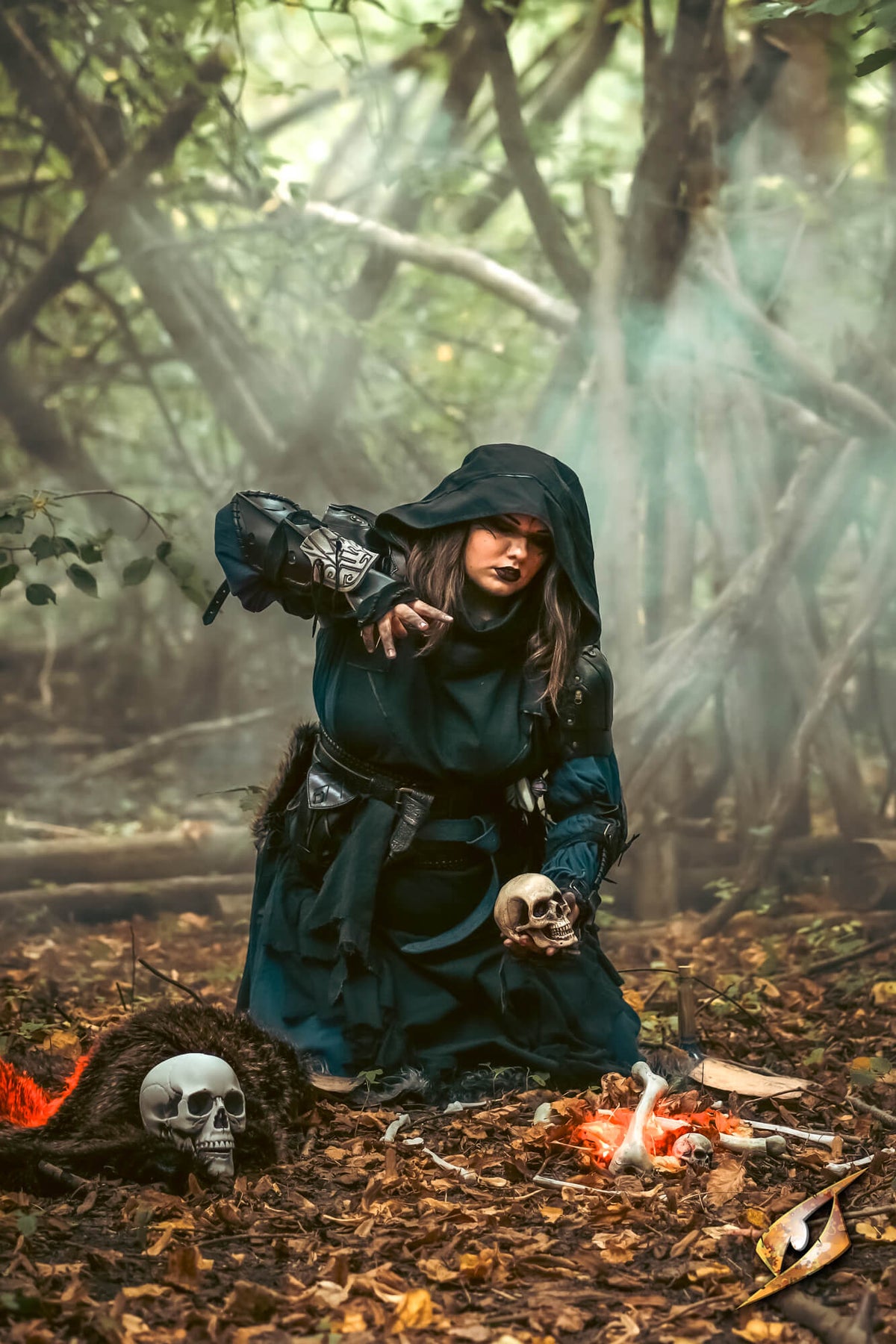 A person in a dark hooded robe kneels in a misty forest setting, holding a skull, with a small fire, another skull, and dark decorative props on the ground. The scene is atmospheric and styled like a gothic or fantasy costume scene.