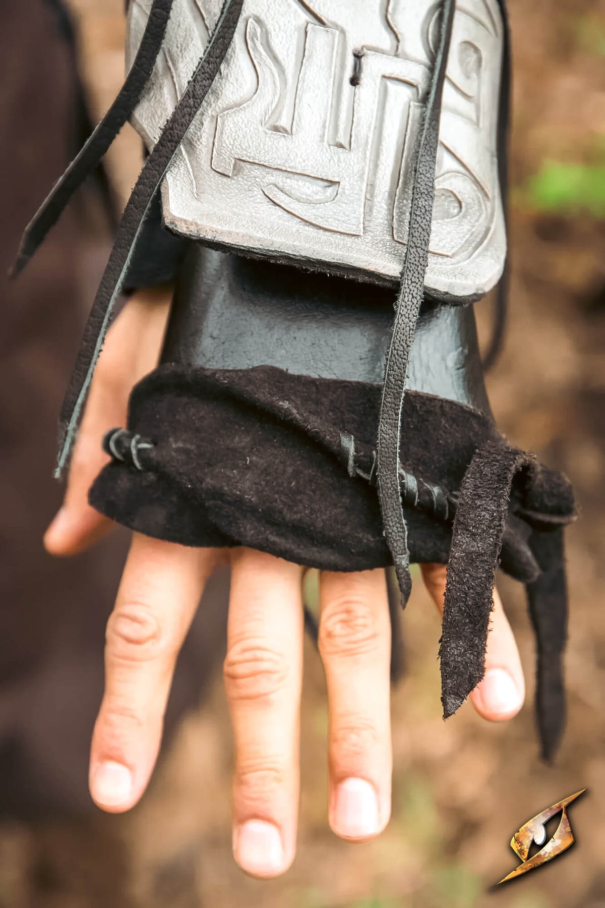 A close-up of a person's hand wearing a dark leather fingerless glove with a silver metal forearm guard featuring an embossed symbol, set against a blurred outdoor background.