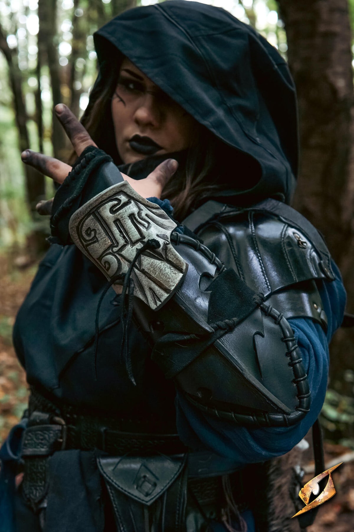 A person wearing a dark hooded fantasy costume with black leather-like armor pieces and a silver-toned arm guard, posed in a forest setting.