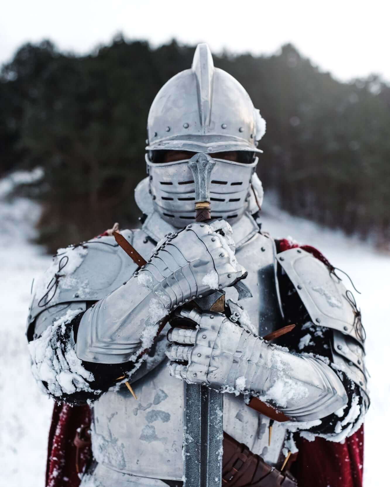 A knight in shining silver armor stands in the snow, holding a sword and wearing a red cape, embodying medieval strength.