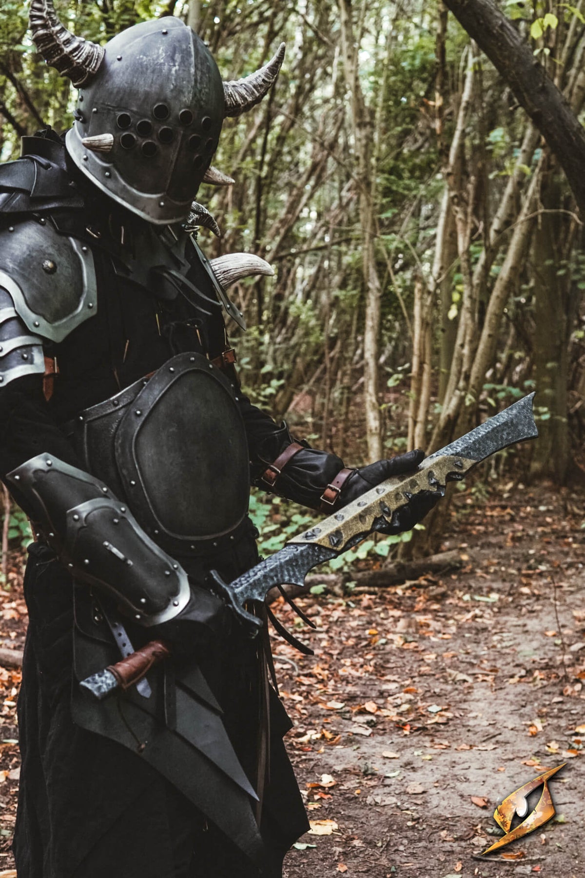A person wearing dark fantasy-style armor with a horned helmet stands in a wooded outdoor setting, holding a large sword. The scene includes a forest path and trees in the background.