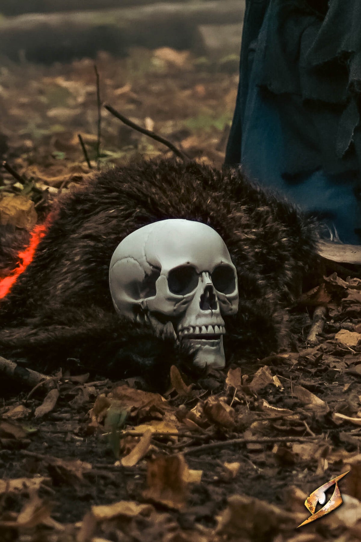 A realistic skull prop resting on dark fur and dry leaves on the ground, with a person's blue pants partially visible in the background.