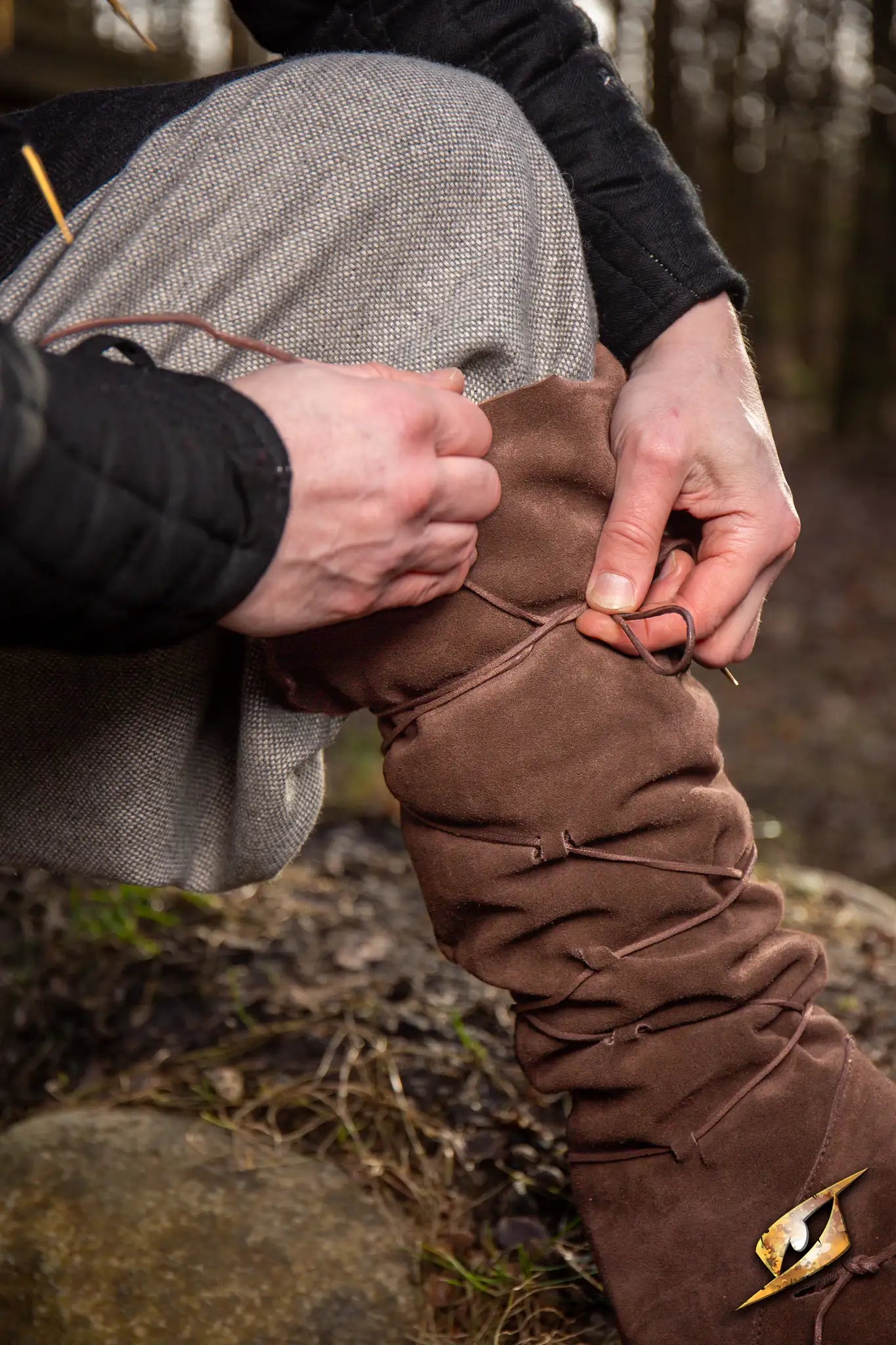 A person tying Theoderic gaiters on their leg in a forest setting, showcasing the product's design and functionality.