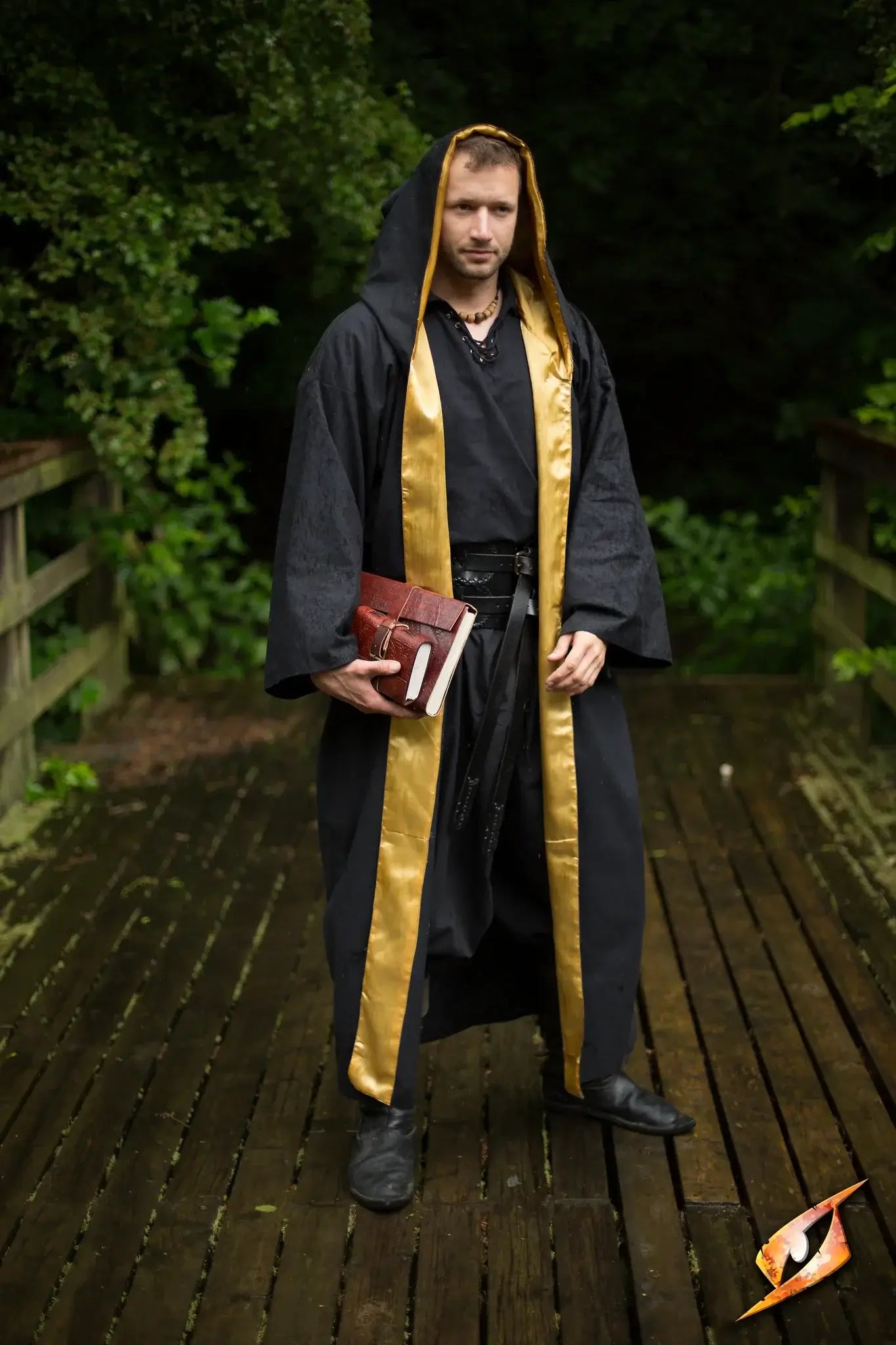 Magician Robe worn by a male magician holding a book, standing on a wooden bridge surrounded by greenery.