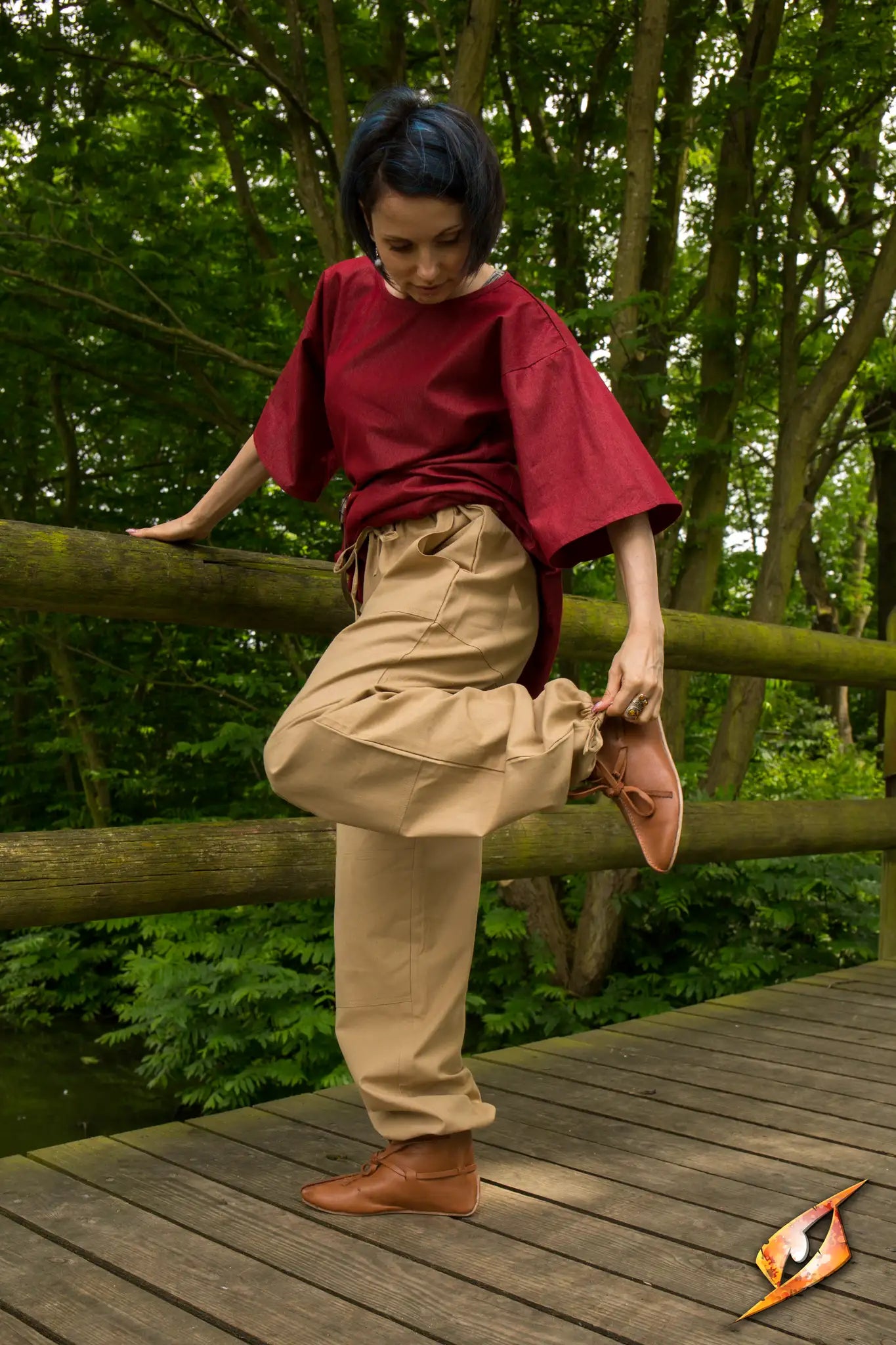 A person wearing Basic Pants and a red top, standing on a wooden bridge surrounded by greenery.