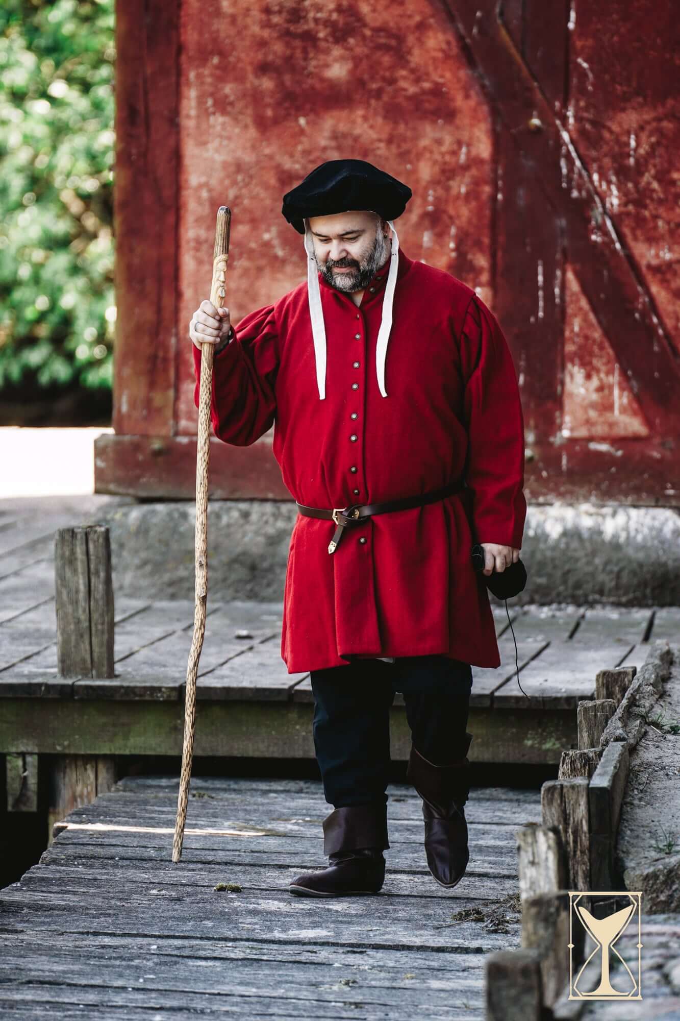 Man wearing Surcoat Lorenzo, a red tunic, walking with a cane by a wooden dock.