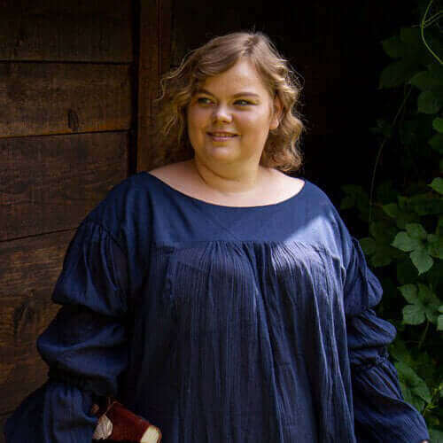 Woman in a dark blue dress standing outside near wooden wall, smiling with natural surroundings.