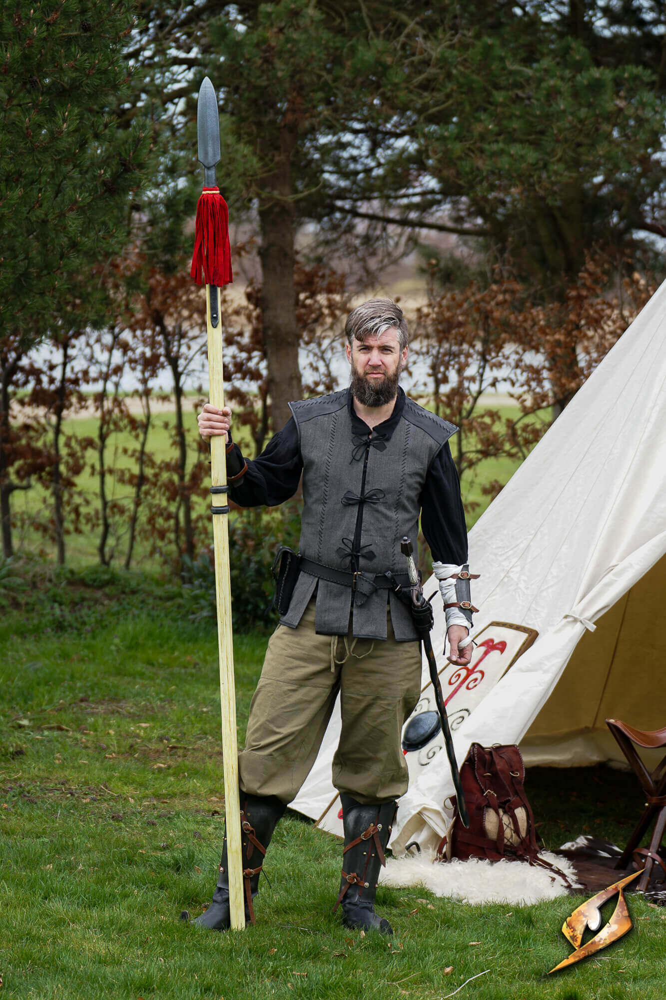 Man in medieval attire holding a spear next to a tent outdoors