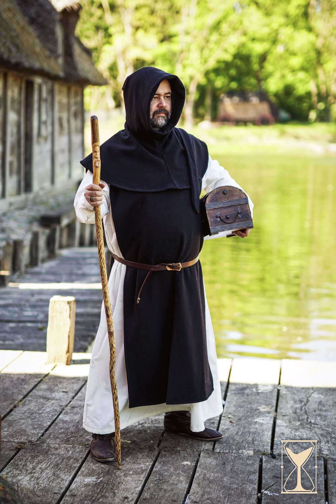A man in a medieval Cyclas Bernard over-tunic, holding a staff and leather pouch by a river.
