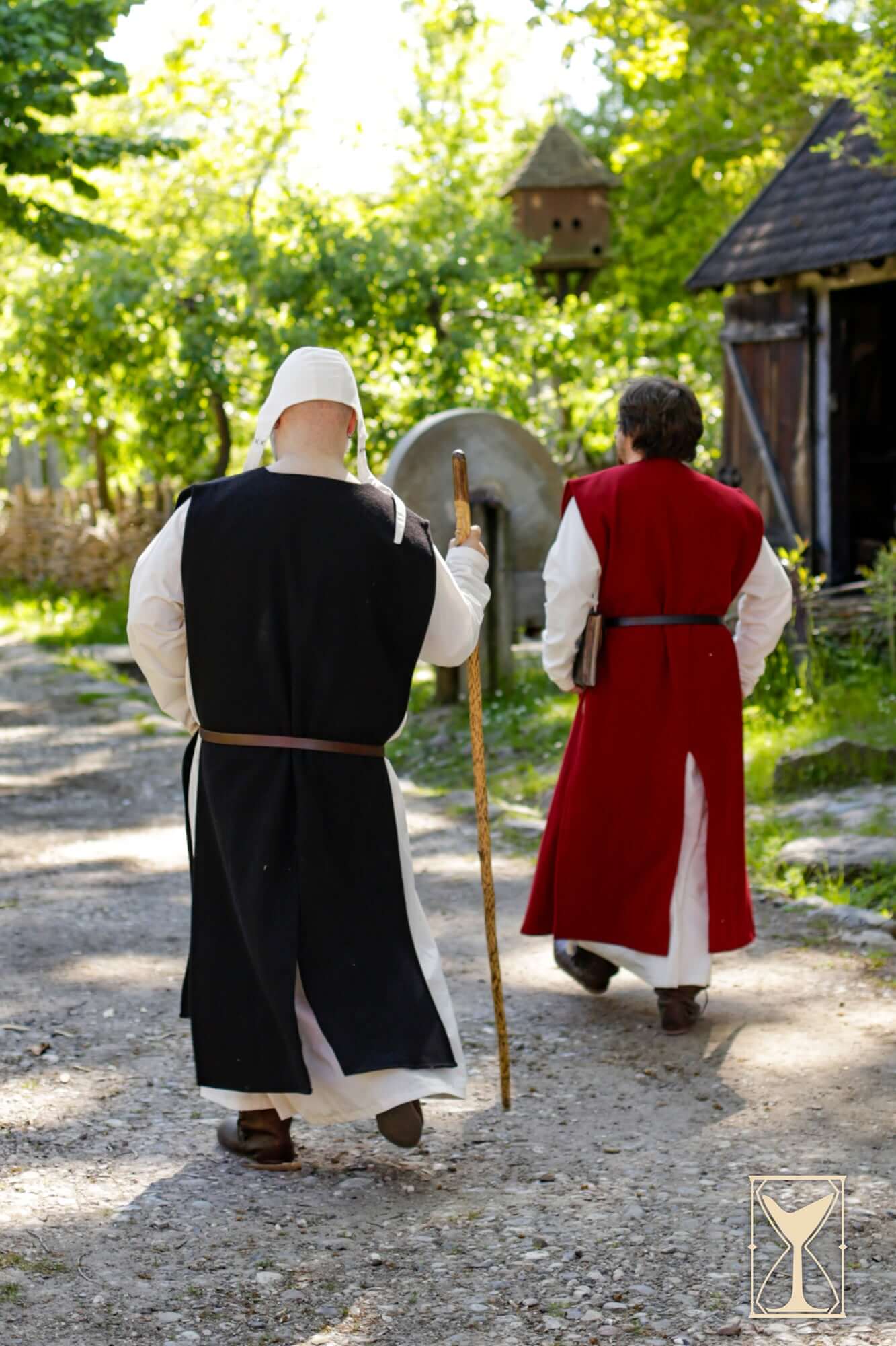 Two individuals in medieval attire wearing the Cyclas Bernard, walking on a pathway in a historical setting.