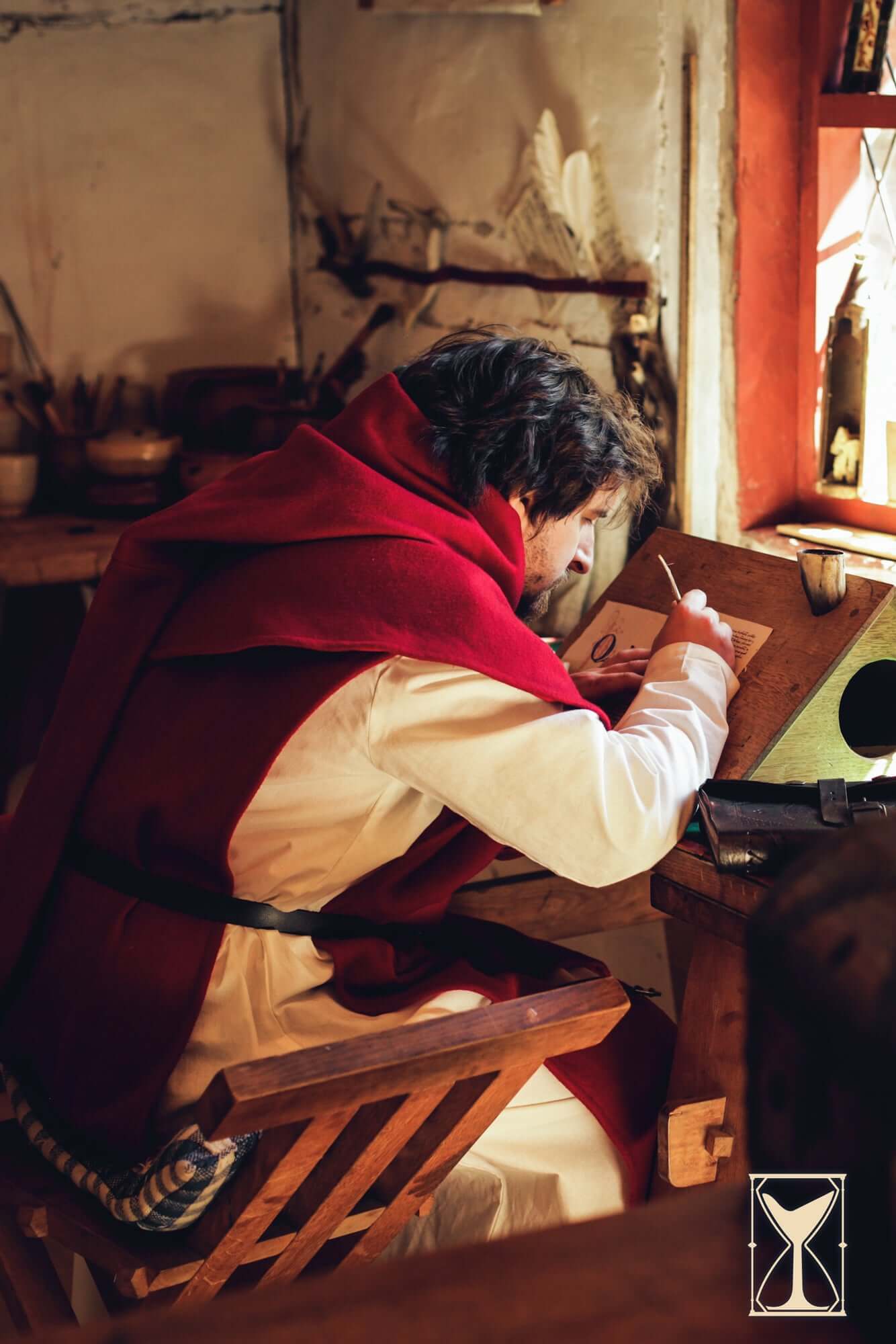 A man in a red Cyclas Bernard over-tunic writing at a wooden desk, capturing the essence of medieval craftsmanship.