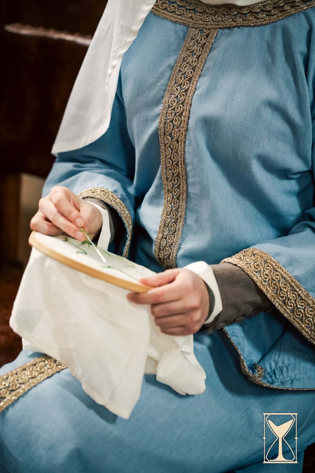 A person wearing the Overtunic Matilde, carefully stitching a decorative pattern on fabric.