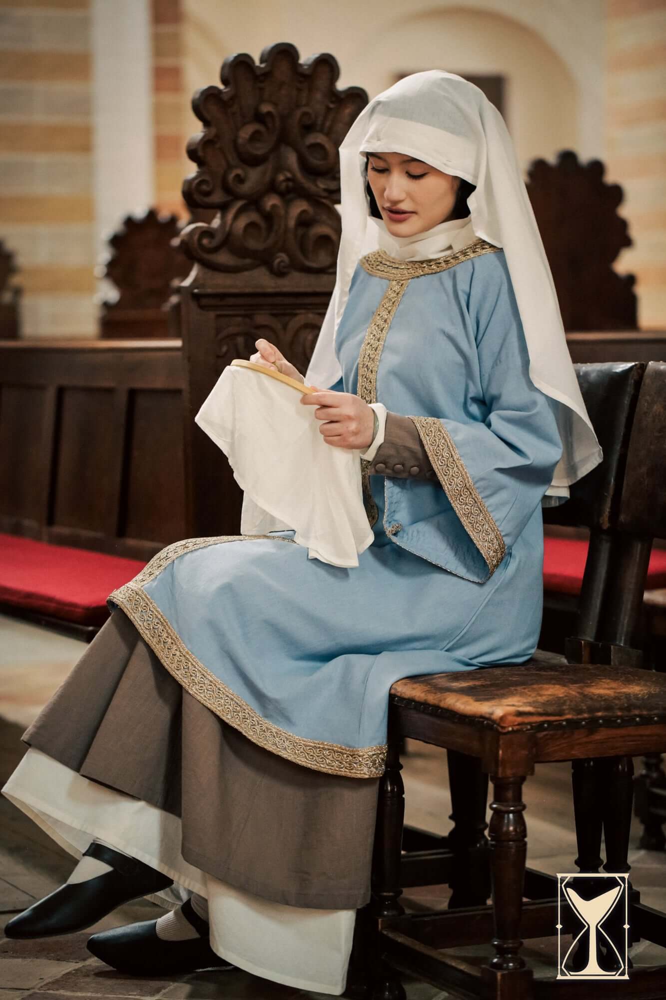 Woman wearing Overtunic Matilde, features historical design and woven patterned trim, seated with fabric.