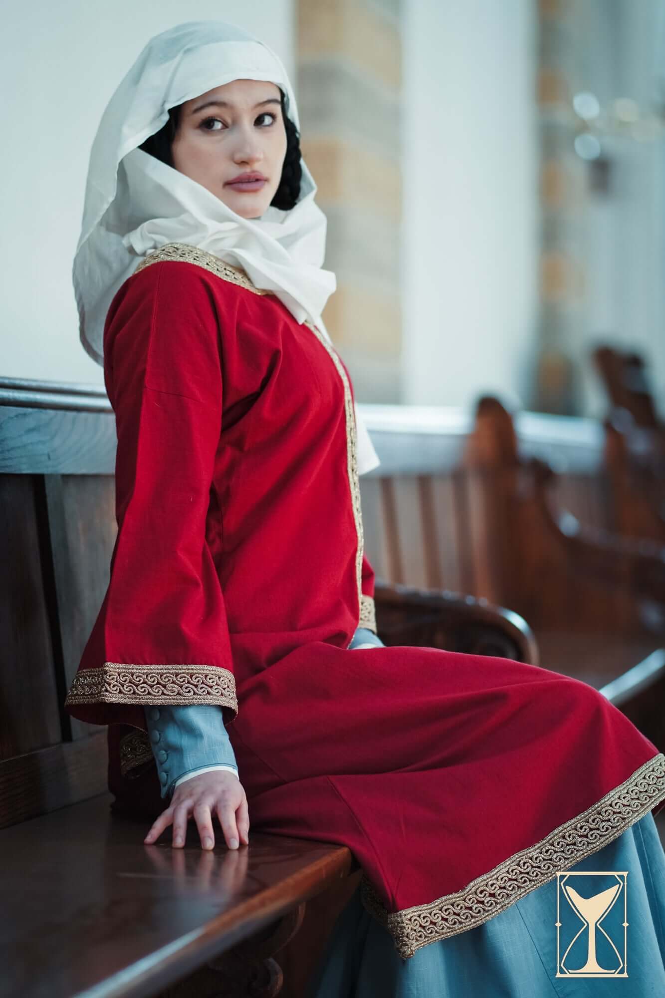 Model wearing the Overtunic Matilde in red with wide sleeves and woven patterned trim, sitting on a wooden bench.