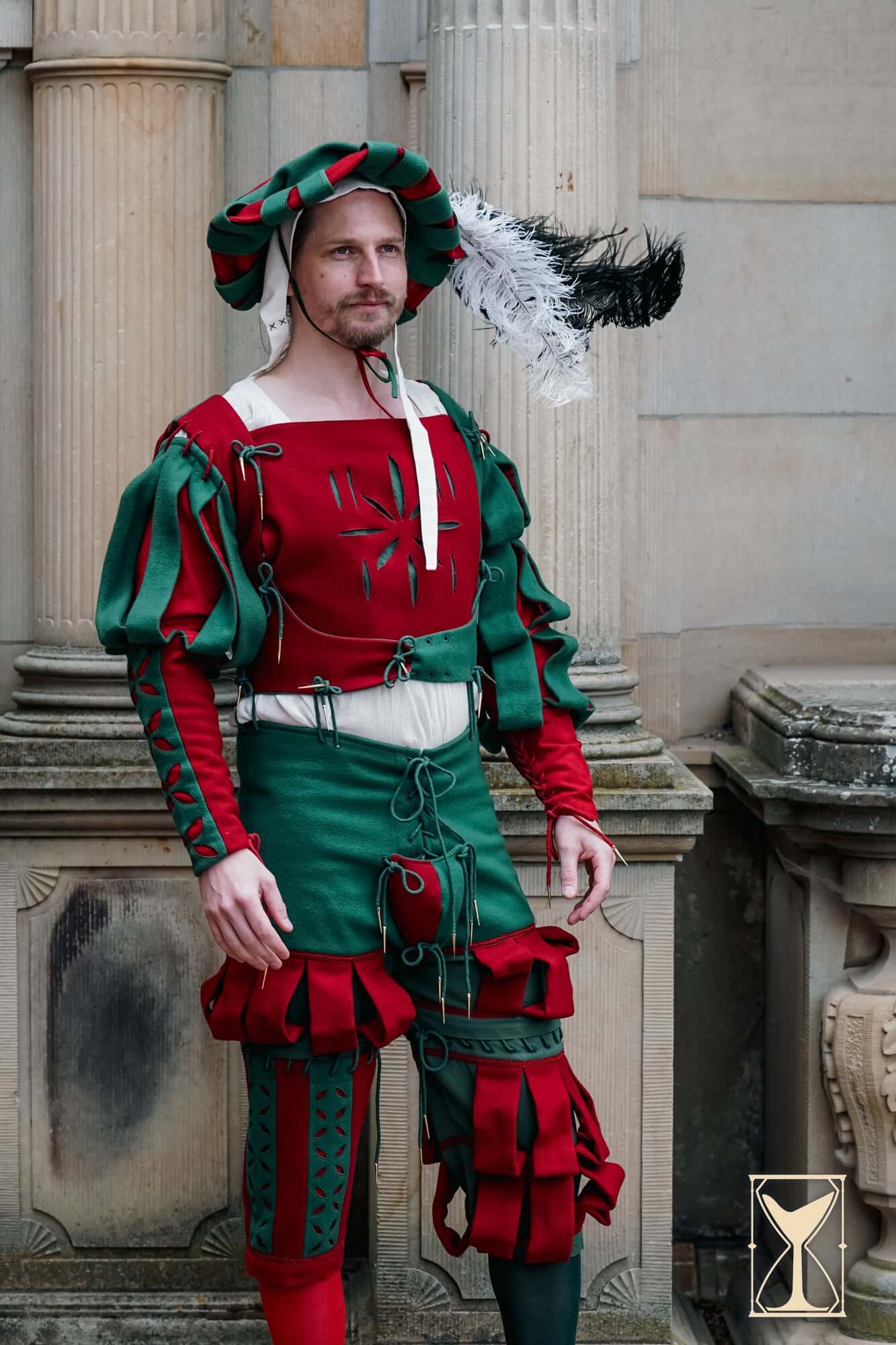 Person in a red and green costume with feathered headdress standing against classical architecture.