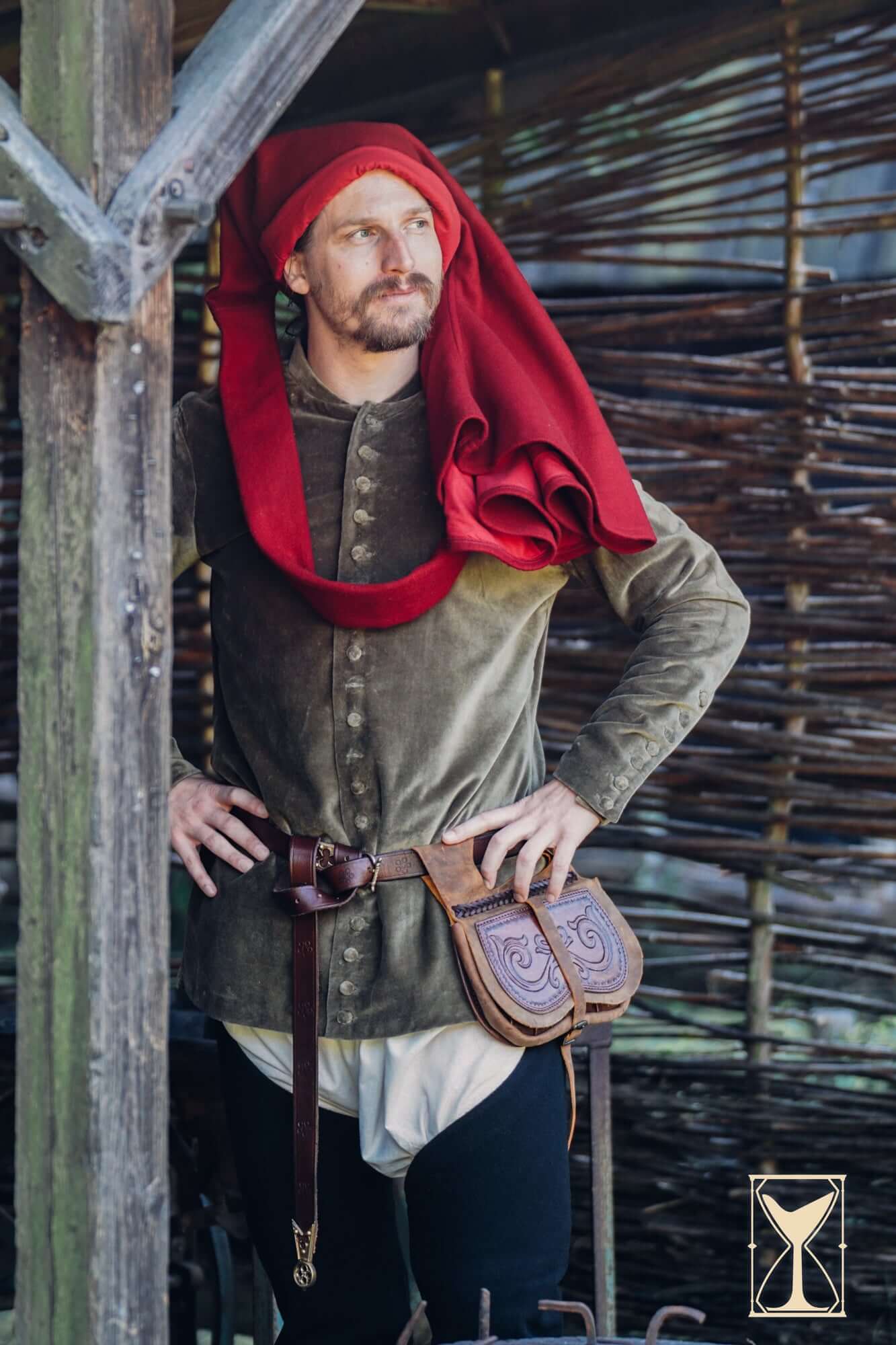 Man in medieval attire with Kidney Bag and red hooded cloak, posing against rustic backdrop.
