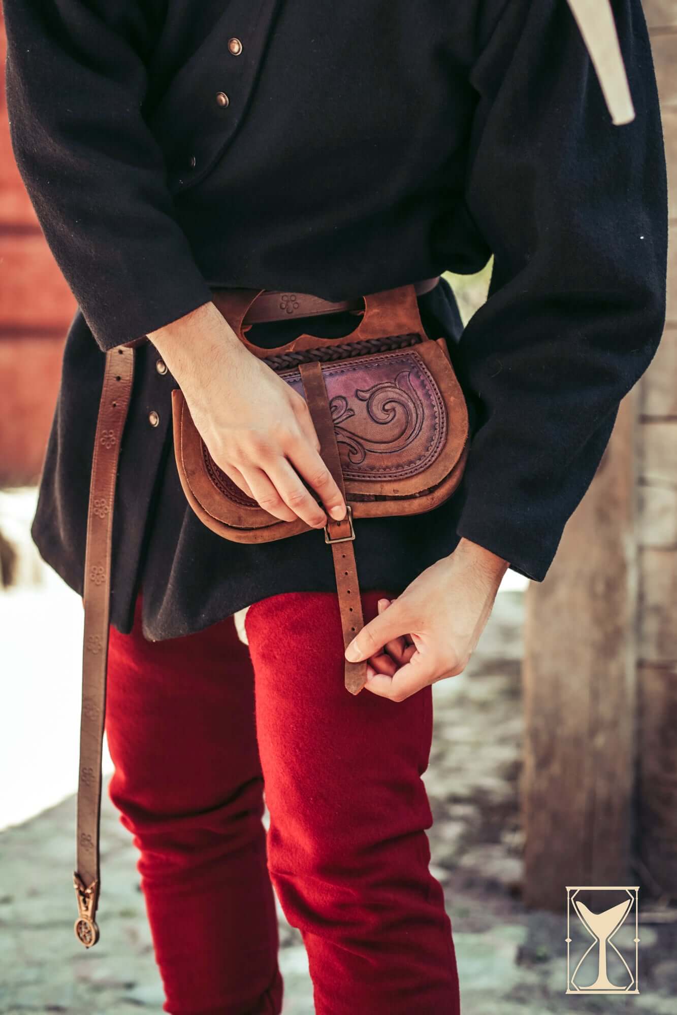 A person adjusting the strap of a Kidney Bag made of brown leather with an engraved design.