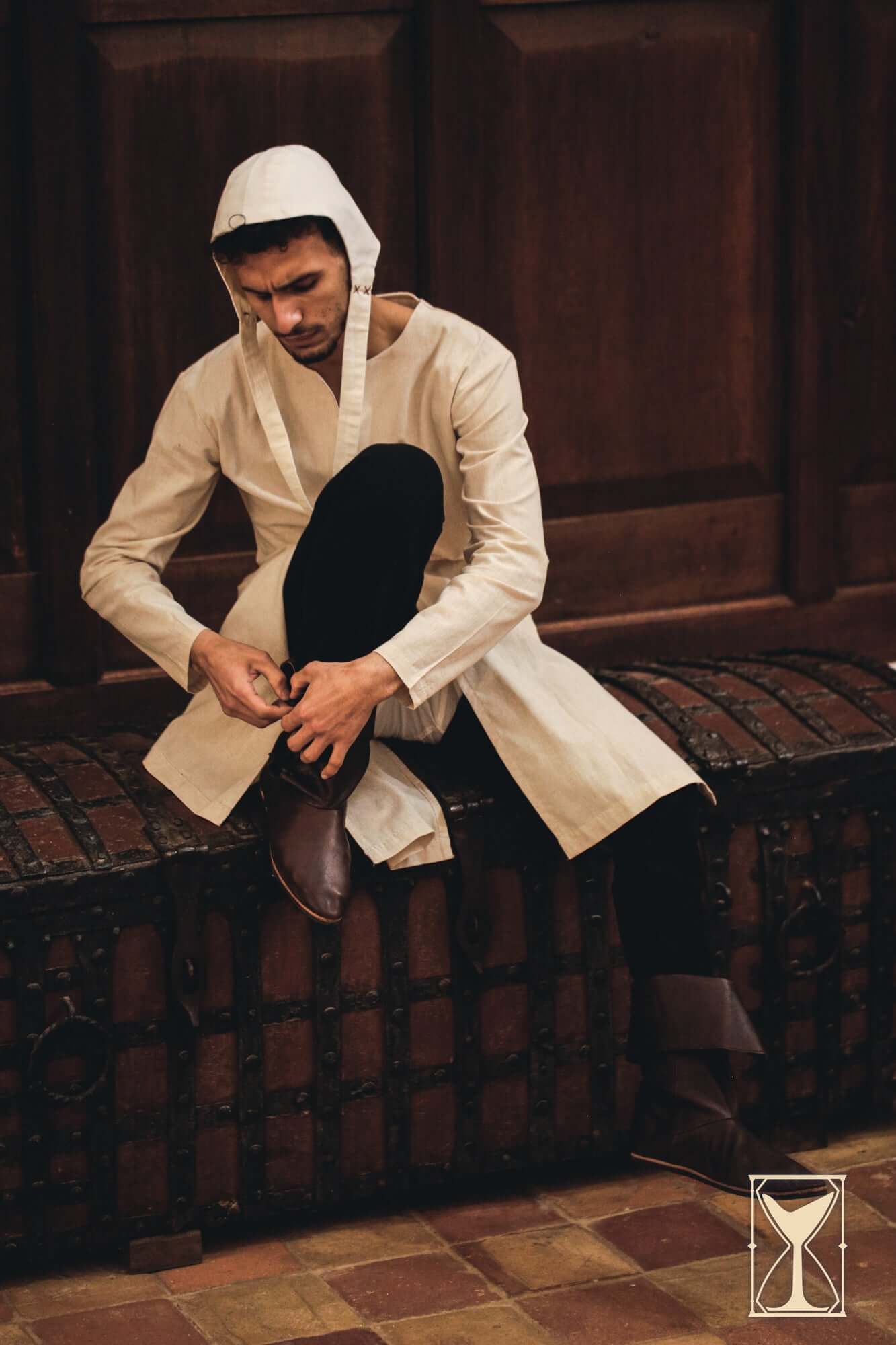 Model wearing a Medieval Riding Tunic while lacing up boots, showcasing its style and functionality.