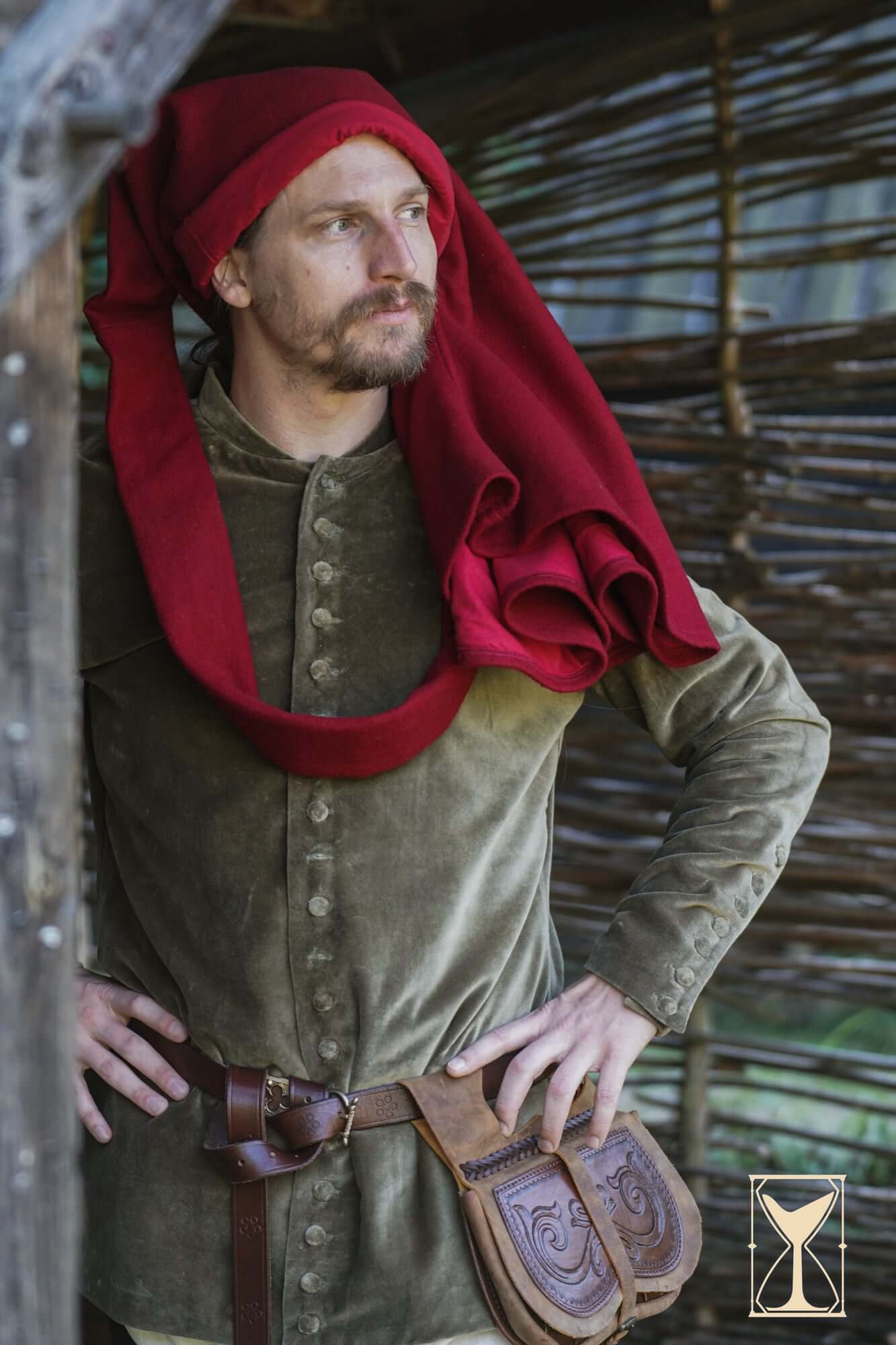 Man wearing Cotehardie Bonifacio with red hood, standing confidently in a rustic setting.