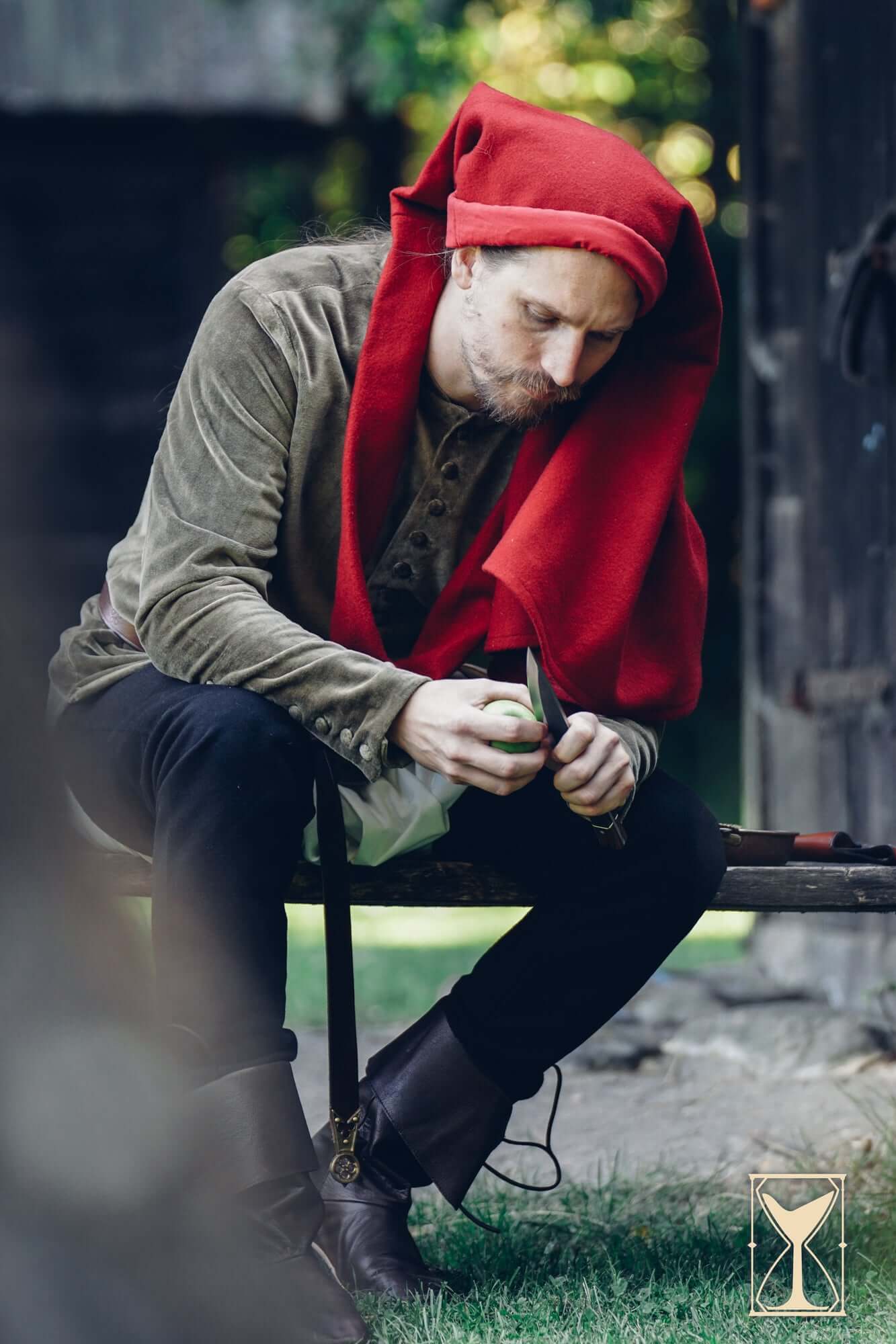 Man wearing Cotehardie Bonifacio with a red hood, sitting outdoors and working with a tool.