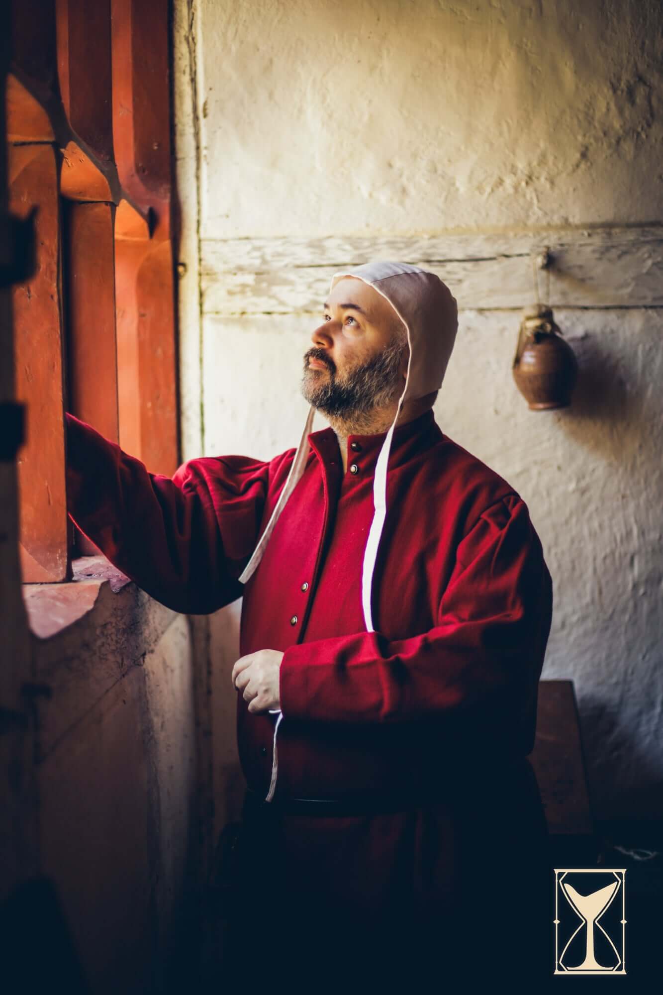 A man in a red outfit wearing a coif 12th century while looking out a window.