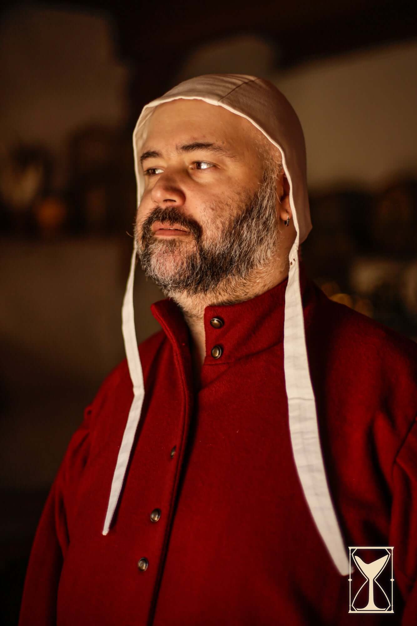 Man wearing a Coif 12th century, a historical cap, in a dimly lit medieval setting.