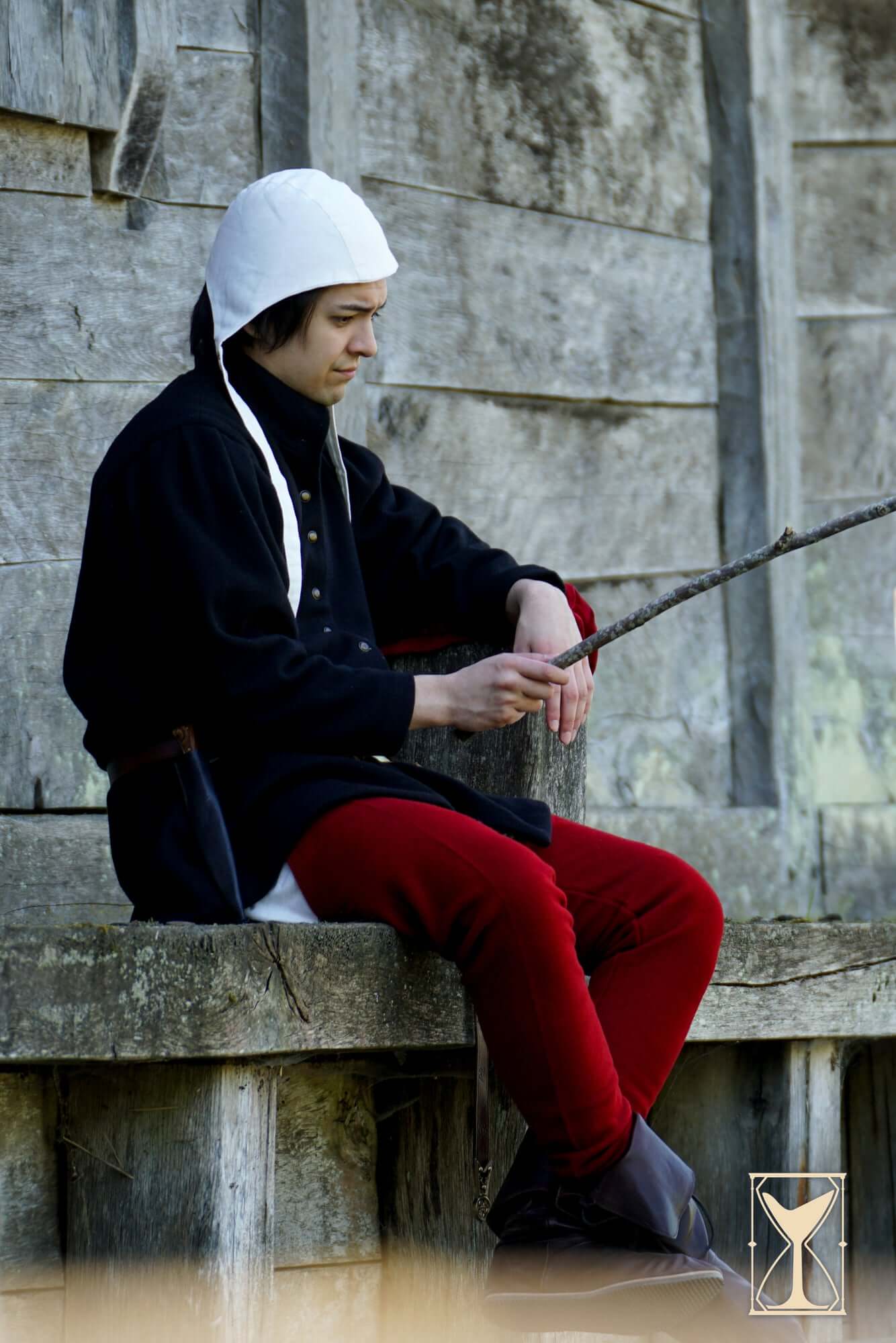 Young person wearing a coif 12th century, seated on a wooden bench, holding a stick outdoors.