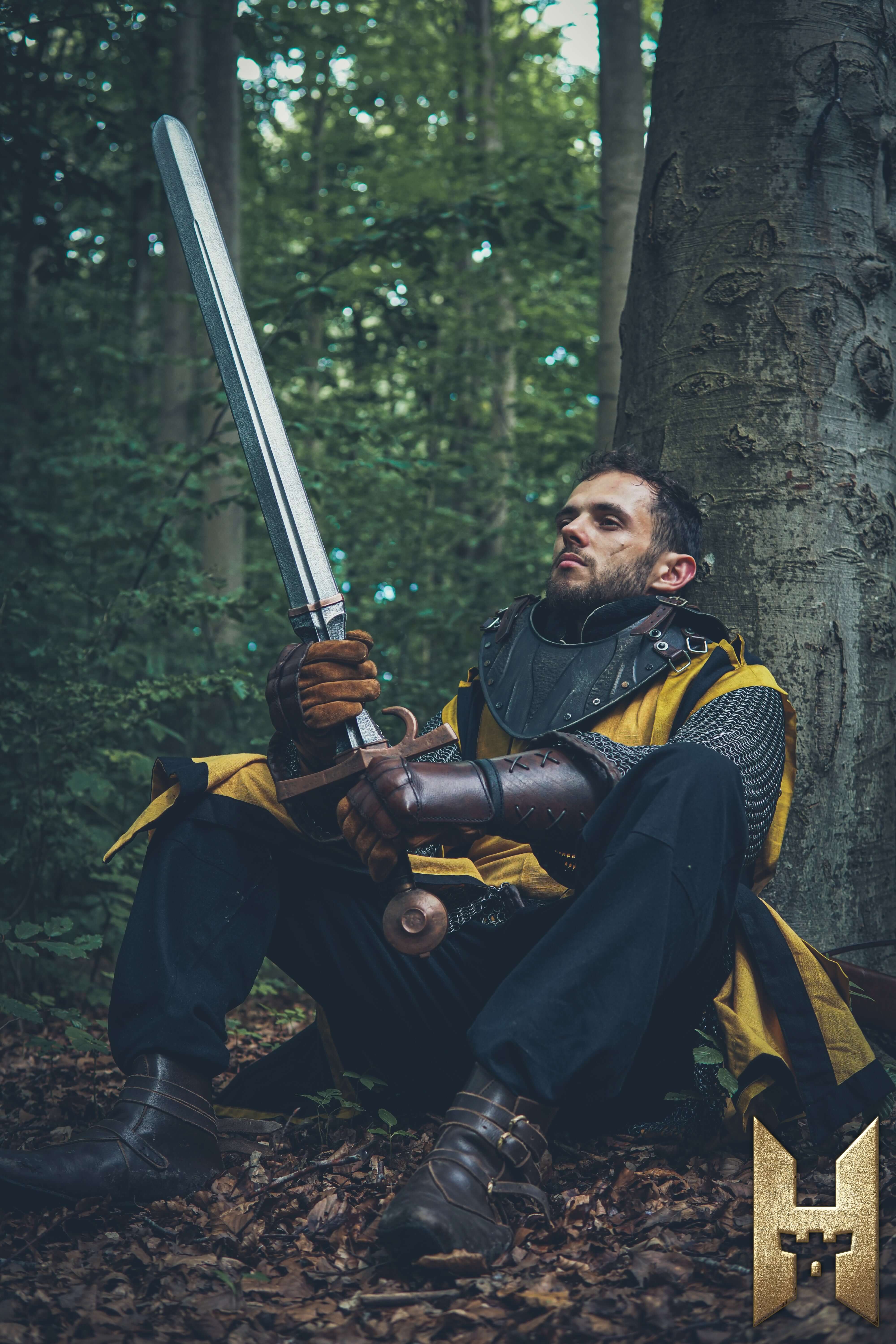 Milanese Sword held by a fighter in medieval armor seated against a tree in a forest.