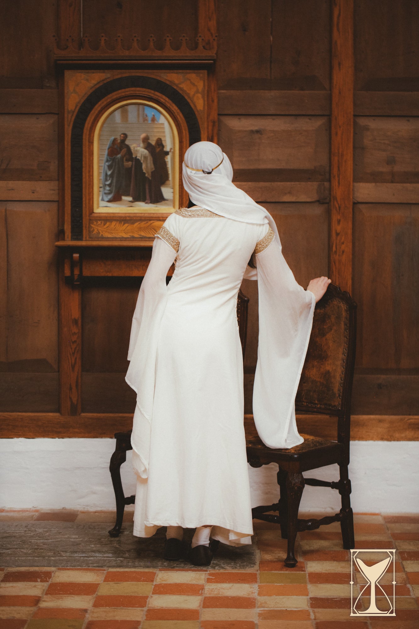 Woman in a Bliaut Noble dress standing beside a chair, showcasing medieval fashion and flowing sleeves.