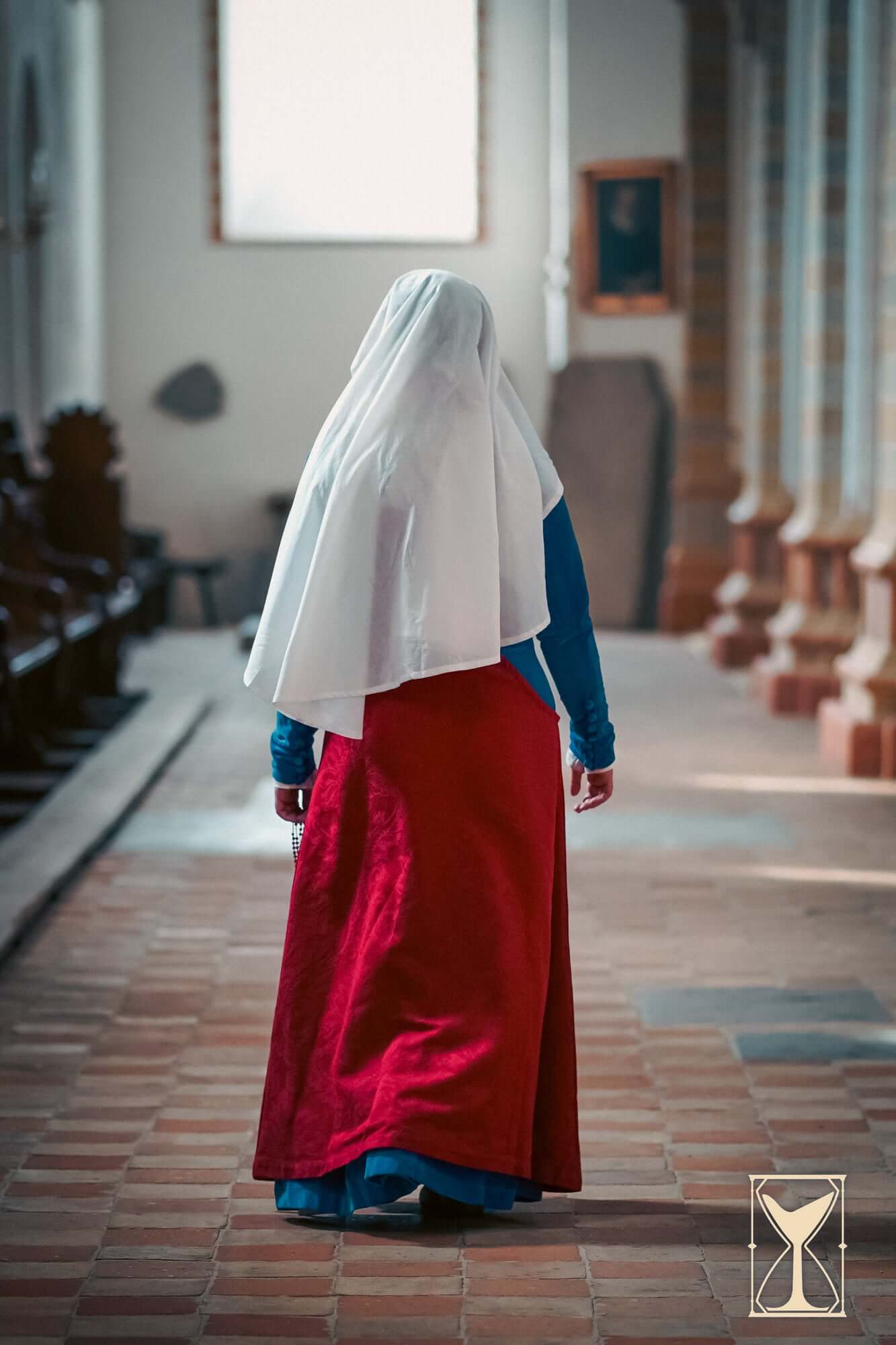 Pellote - Medieval Dress worn by a woman in a historic church setting, featuring a blue bodice and flowing red skirt.