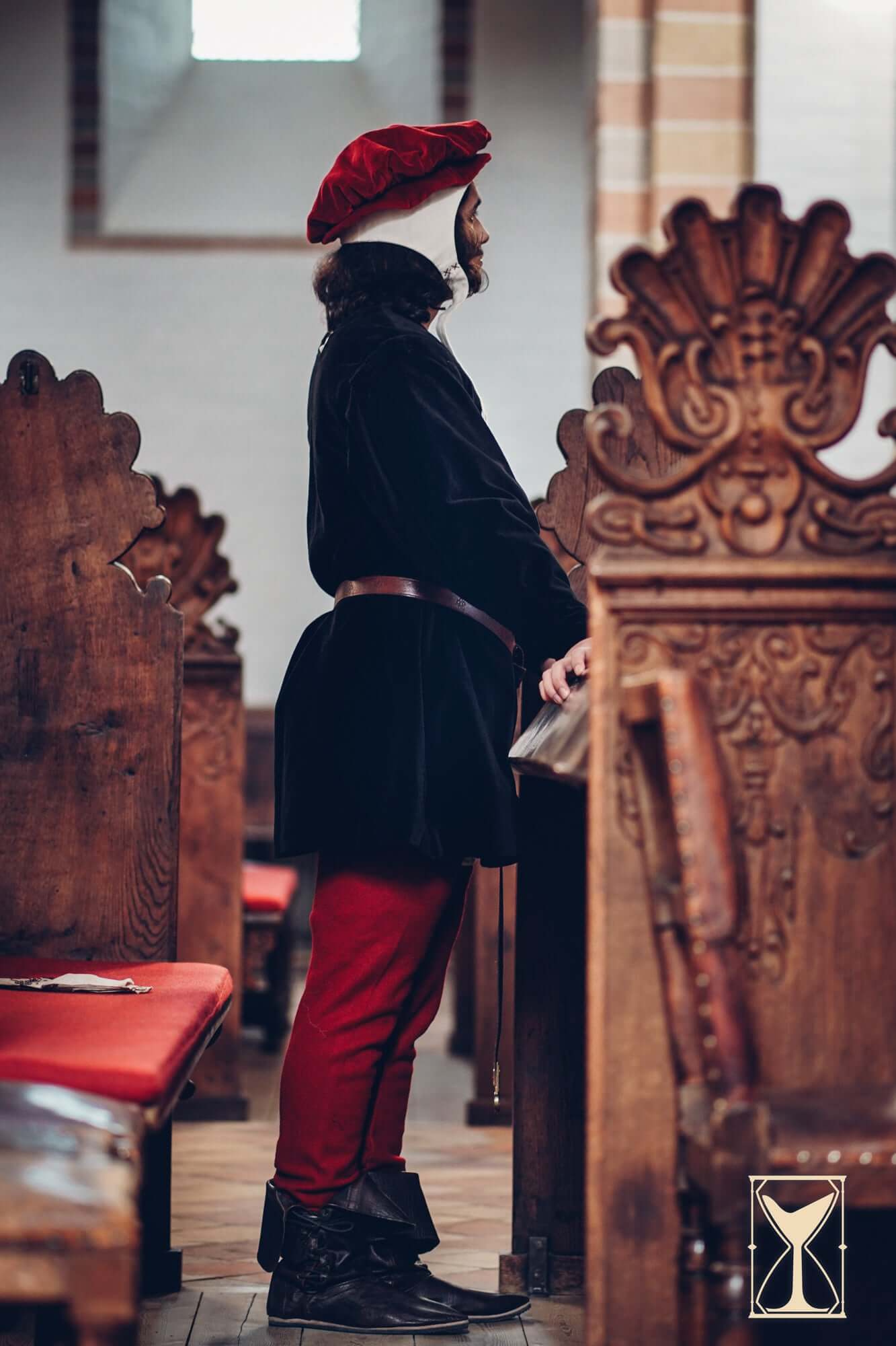 Man wearing a Velvet Surcoat and red pants stands in a medieval-style church among wooden pews.