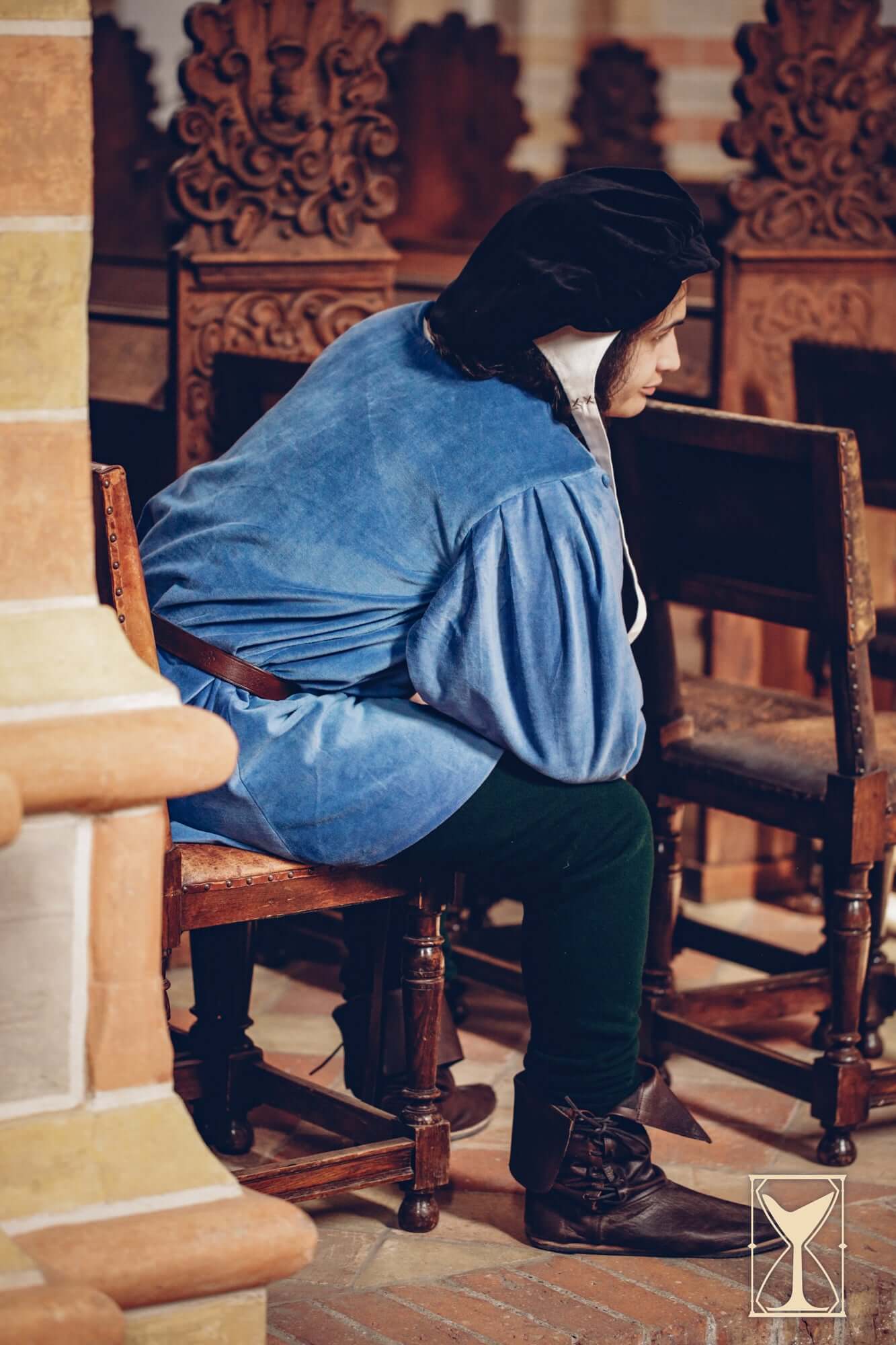 Person seated wearing a Velvet Surcoat, showcasing its rich texture and medieval design in a historical setting.