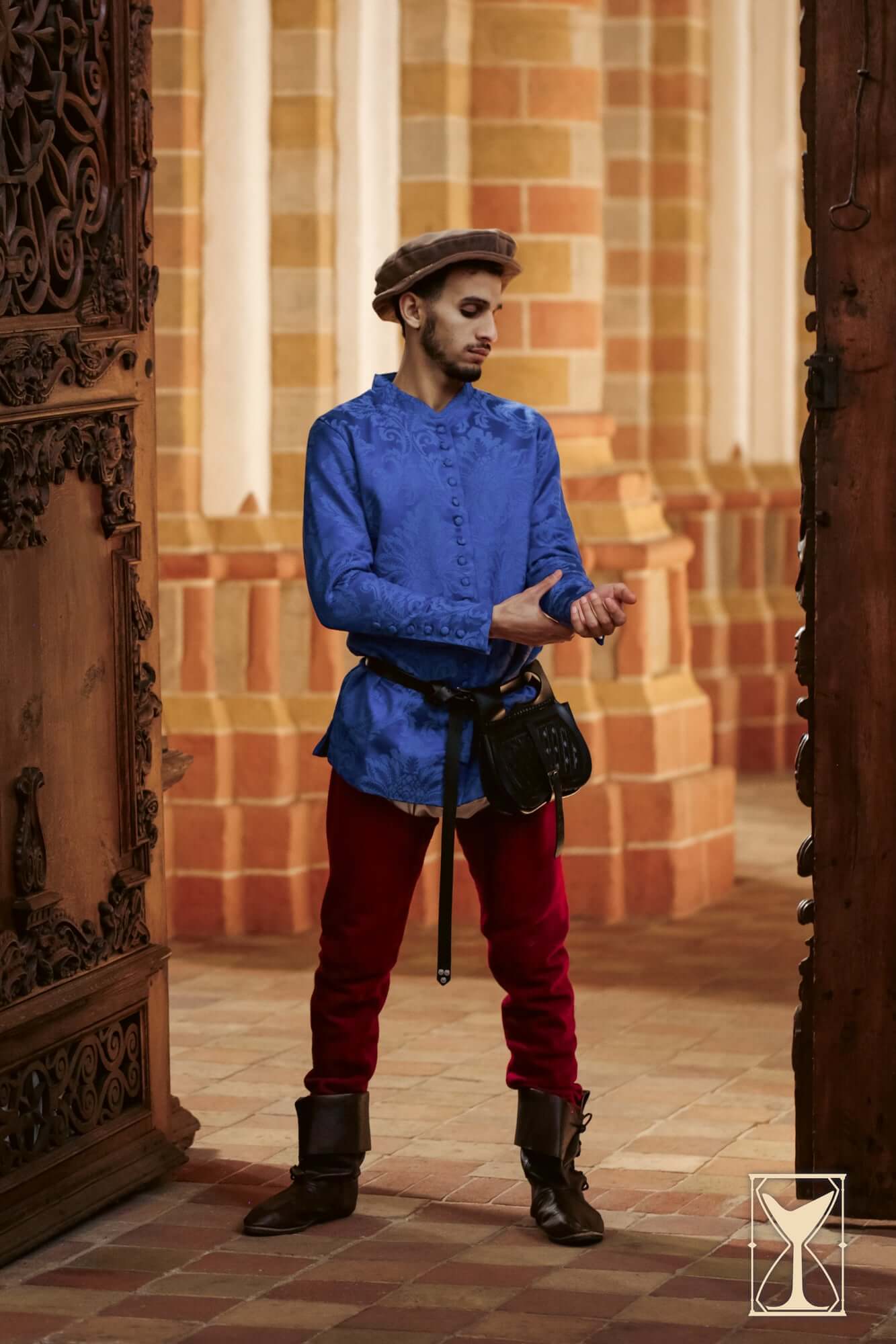 Cotehardie Rodrigo worn by a model, featuring a blue brocade jacket and red trousers, set in a medieval-style interior.