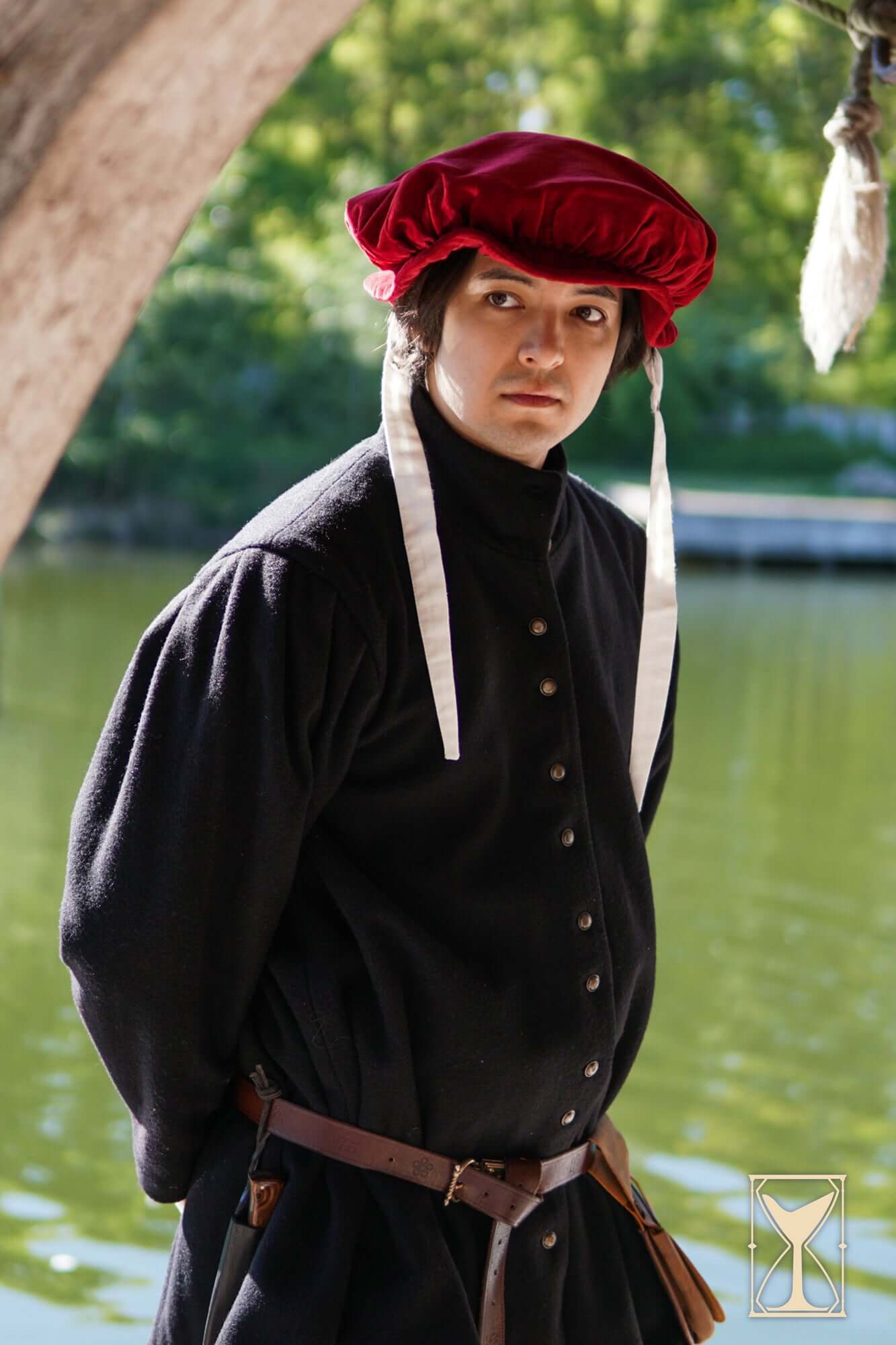 Man wearing a Beret Francisco, a velvet headpiece reflecting 15th and 16th-century fashion, by a serene water backdrop.