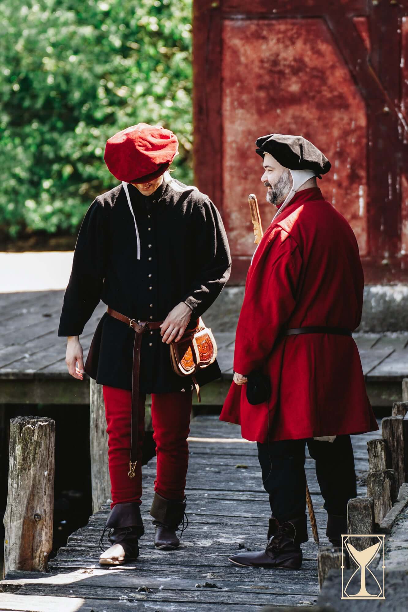 Two men in historical clothing, one wearing a red Beret Francisco, walking on a wooden dock.
