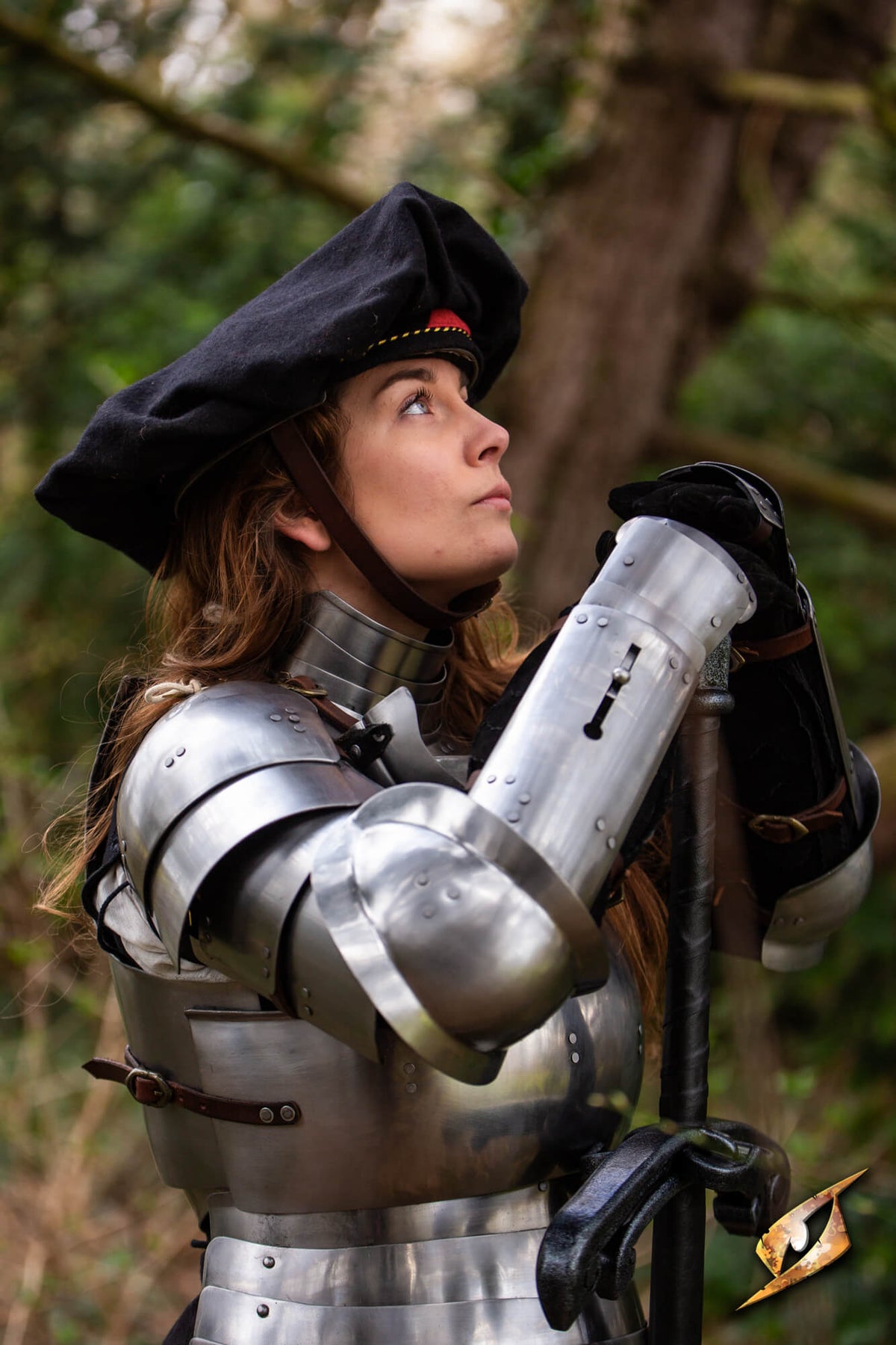 A woman in medieval-style steel armor and a black hat stands in a forest, holding a long pole or staff. The background is a blurred natural woodland setting.