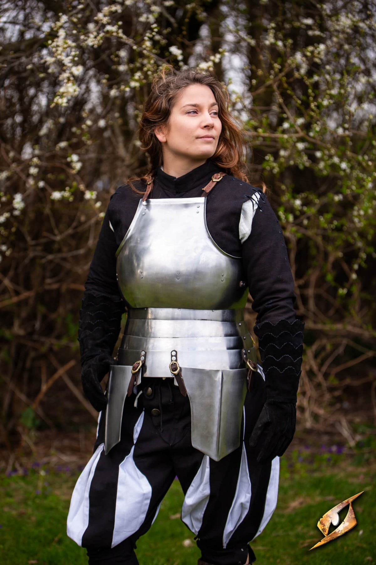 A woman wearing a medieval knight or crusader-style armor costume stands outdoors in front of flowering trees. The costume includes a silver breastplate, a metal waist guard, black sleeves with gold accents, and black-and-white puffed pants.
