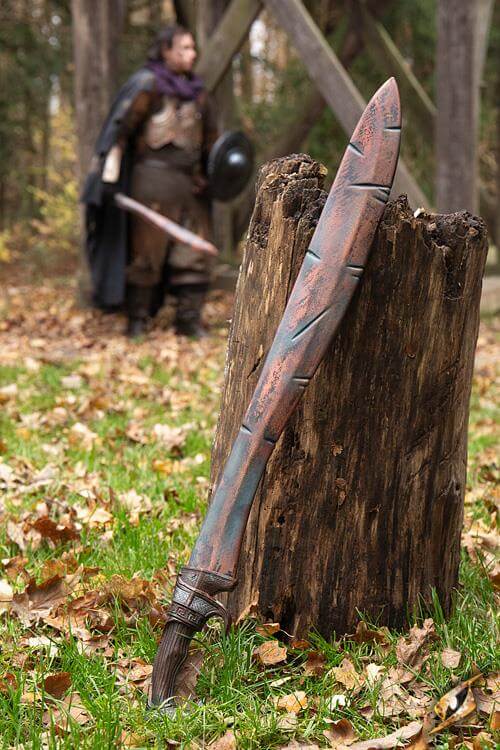 Battleworn Falcata resting on a stump with a warrior in the background, showcasing its handcrafted design.