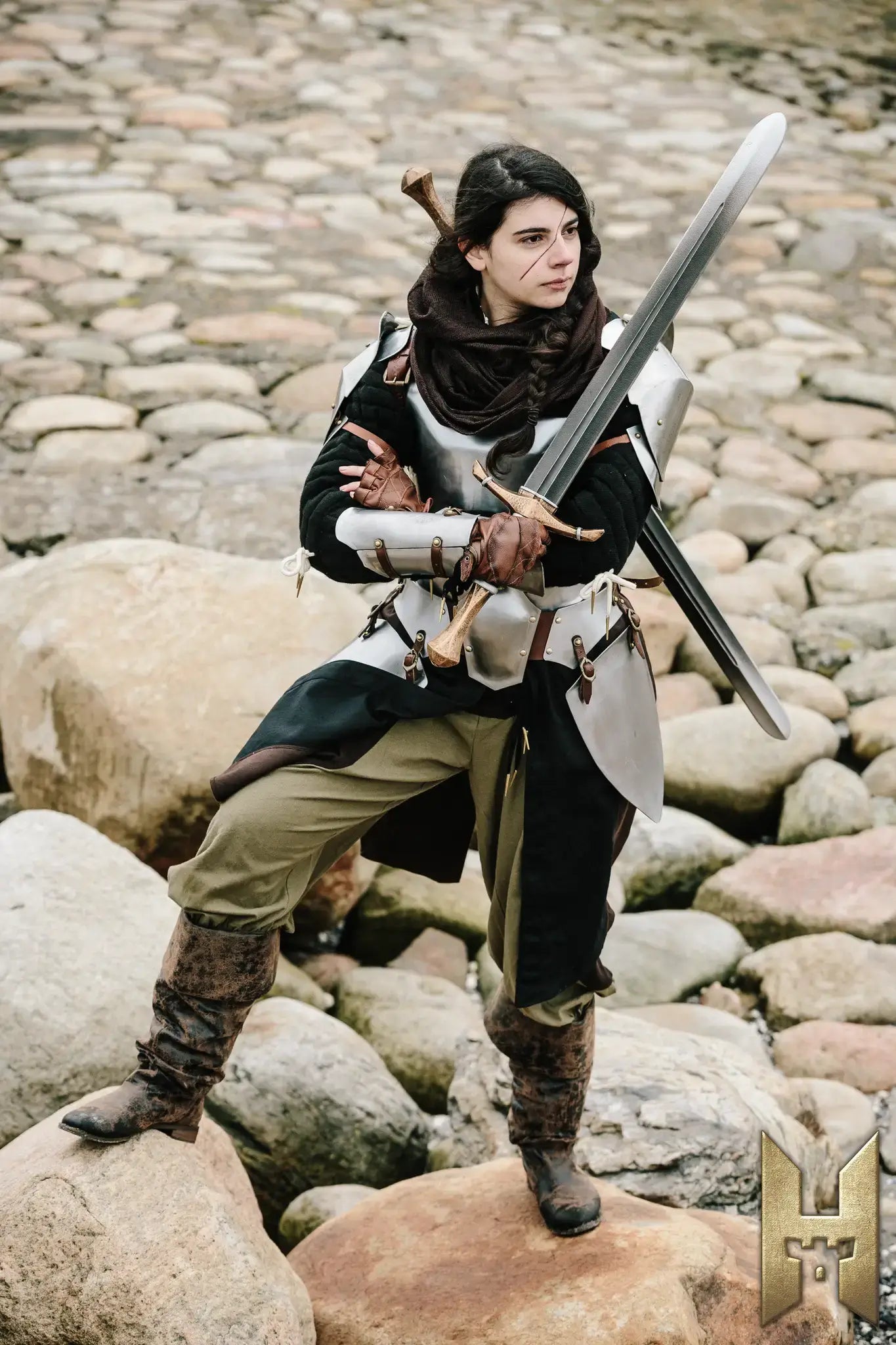 Woman in armor holding a Bastard Sword, posing confidently on rocky terrain.