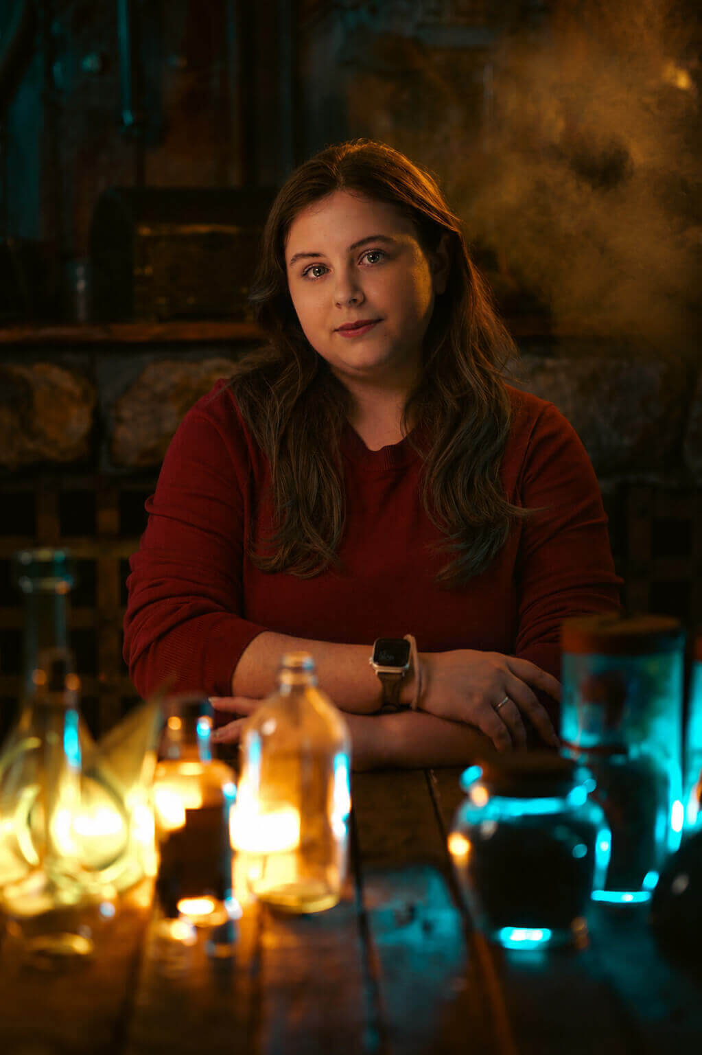 A woman with long brown hair sits at a rustic wooden table in a dimly lit stone interior, wearing a dark red long-sleeve top. Glowing glass bottles and jars sit on the table in warm yellow light, with cool blue light reflections on the right side of the scene.