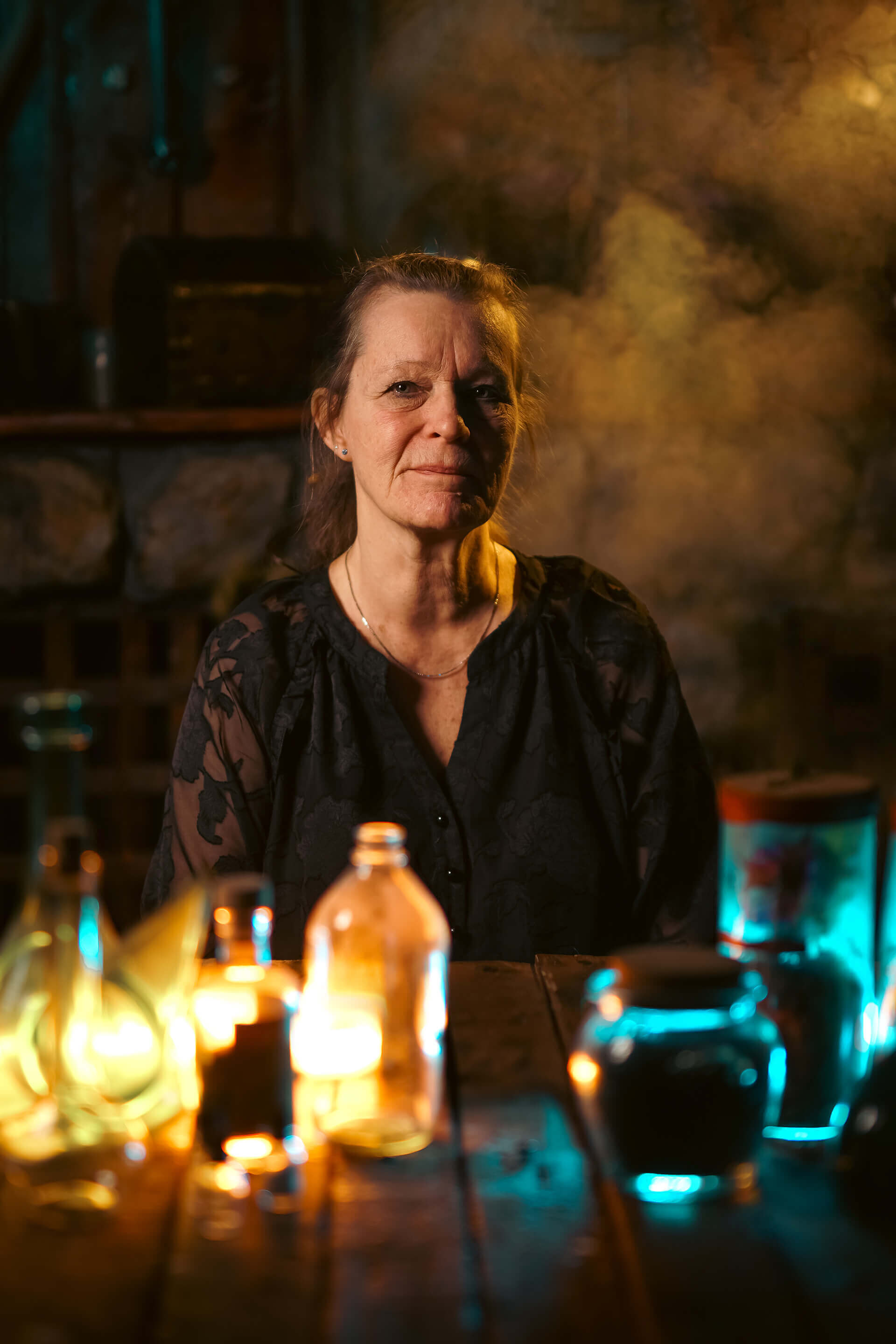 A woman sitting at a table surrounded by colorful bottles and smoke, creating a mystical atmosphere.