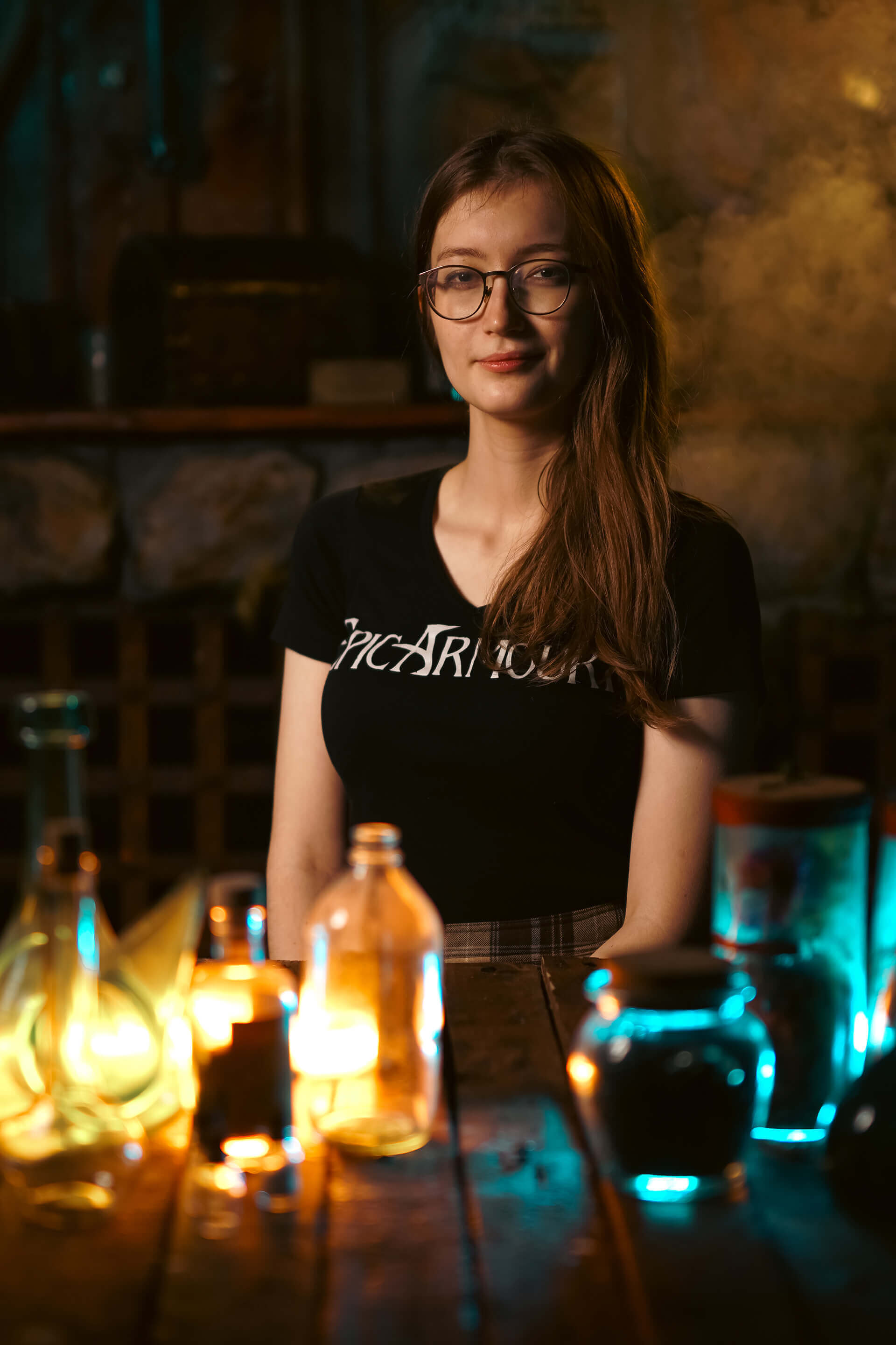 Young woman with glasses sitting at a table surrounded by various glowing bottles in a dimly lit setting.