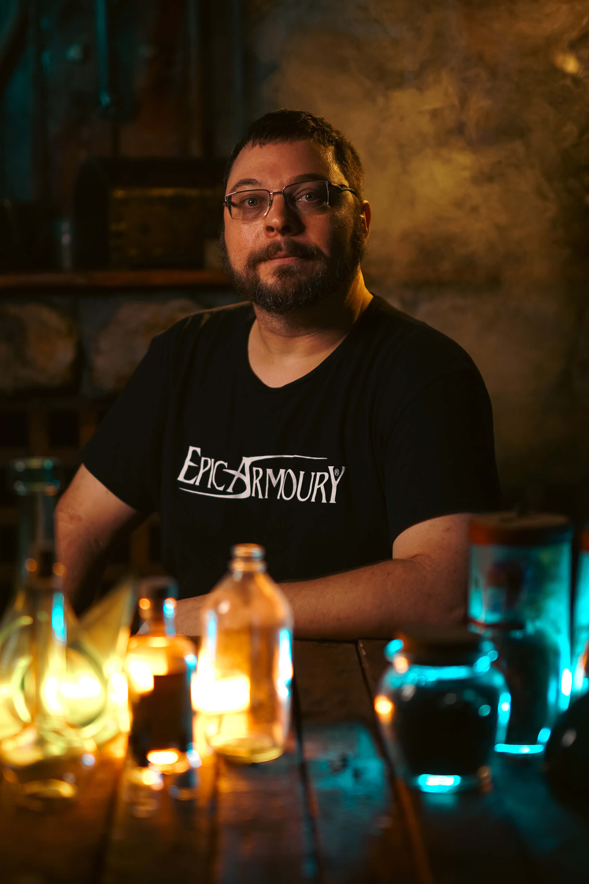 Man sitting at a table with illuminated bottles, wearing an Epic Armoury shirt in a dimly lit setting.