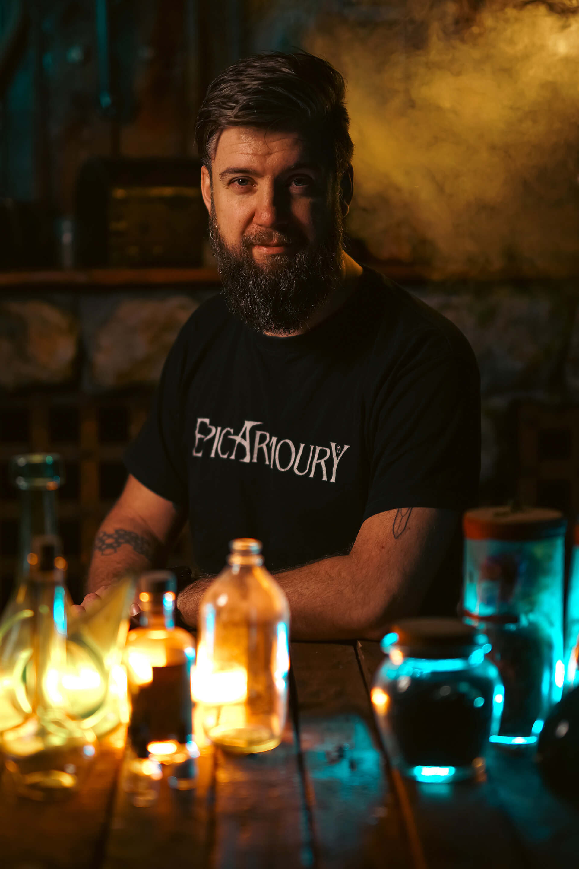 Portrait of a man with a beard in a dimly lit setting, surrounded by colorful glass bottles and wearing an Epic Artoury shirt.