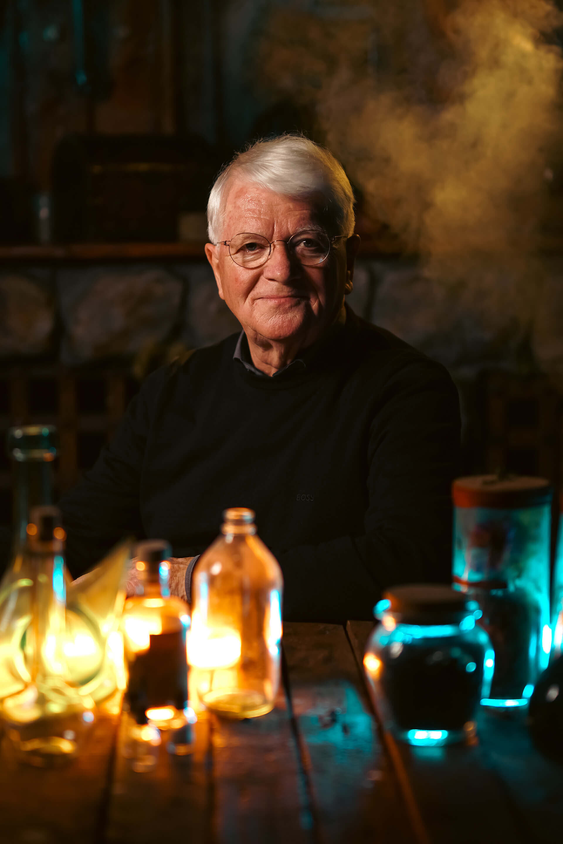 A smiling elderly man with glasses sits at a wooden table surrounded by glowing bottles and jars.