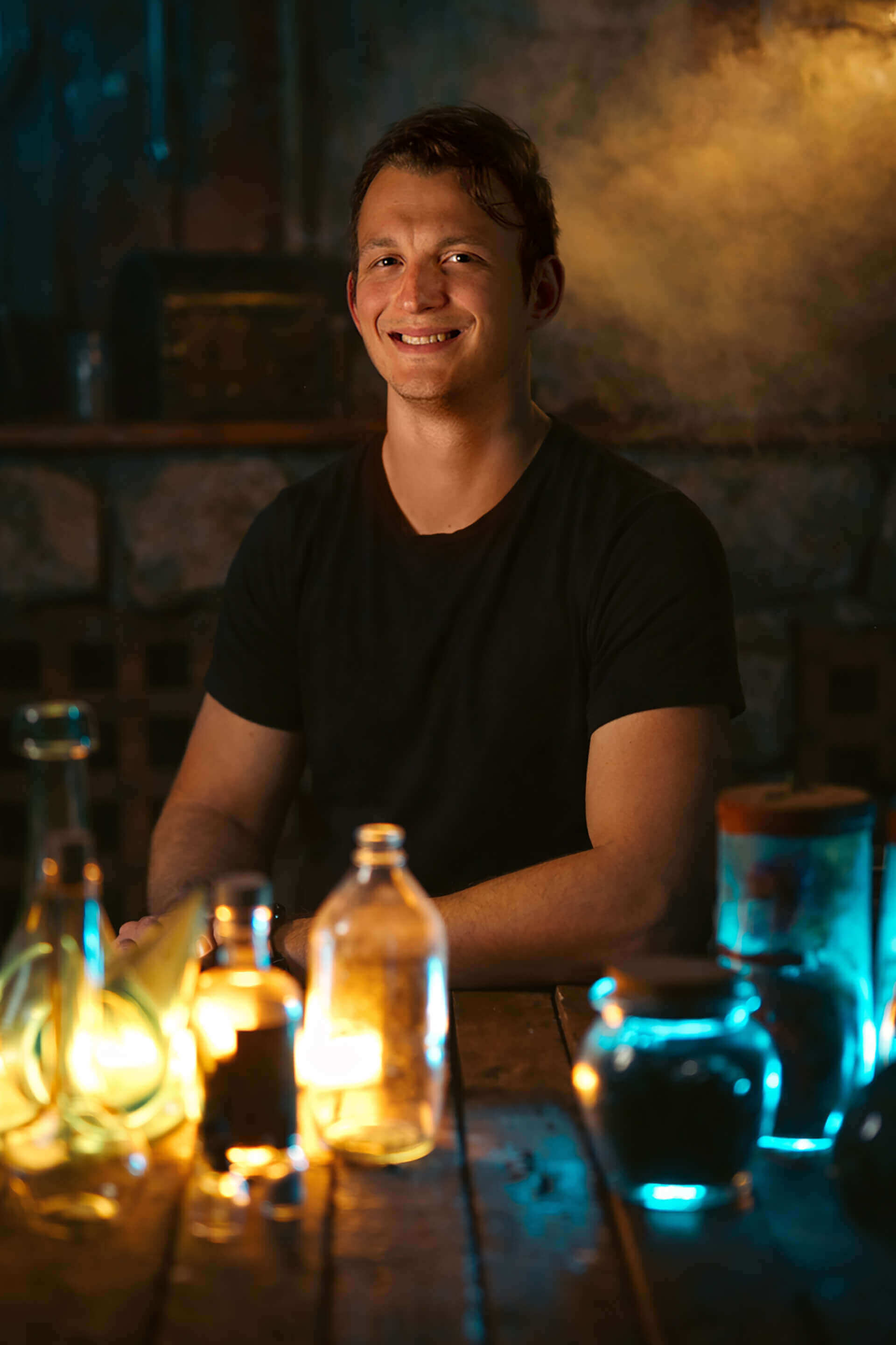 Smiling man in a black shirt sitting at a wooden table with colorful glowing bottles and jars.